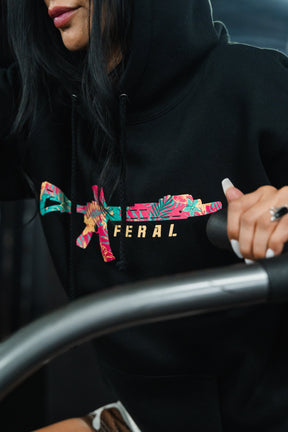 A person wears the black Feral - Premium Hoodie, featuring a rifle graphic in a floral pattern with the word FERAL in gold, capturing a sense of radical authenticity.