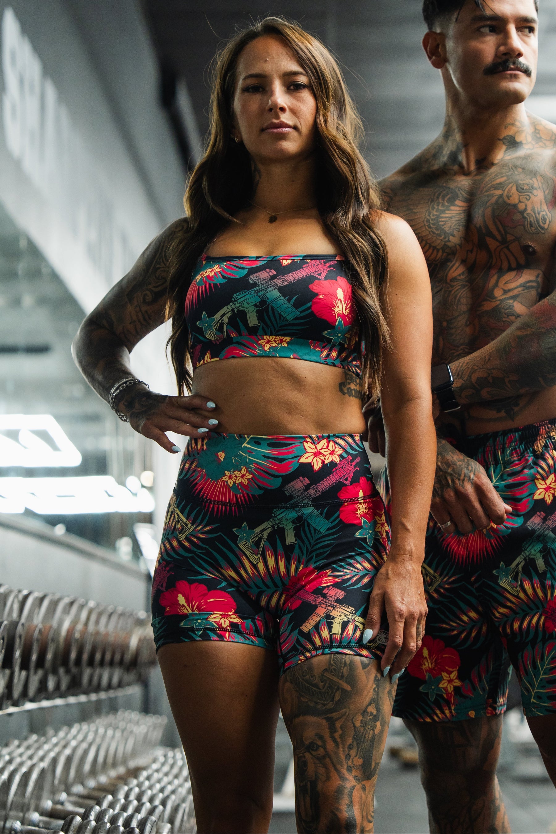 A tattooed woman and man wear matching floral Tango Charlie Apparel outfits, including Women's High-Waisted Seamless Front Compression Training Shorts | Tropical Assassin, standing confidently in a gym near dumbbells.