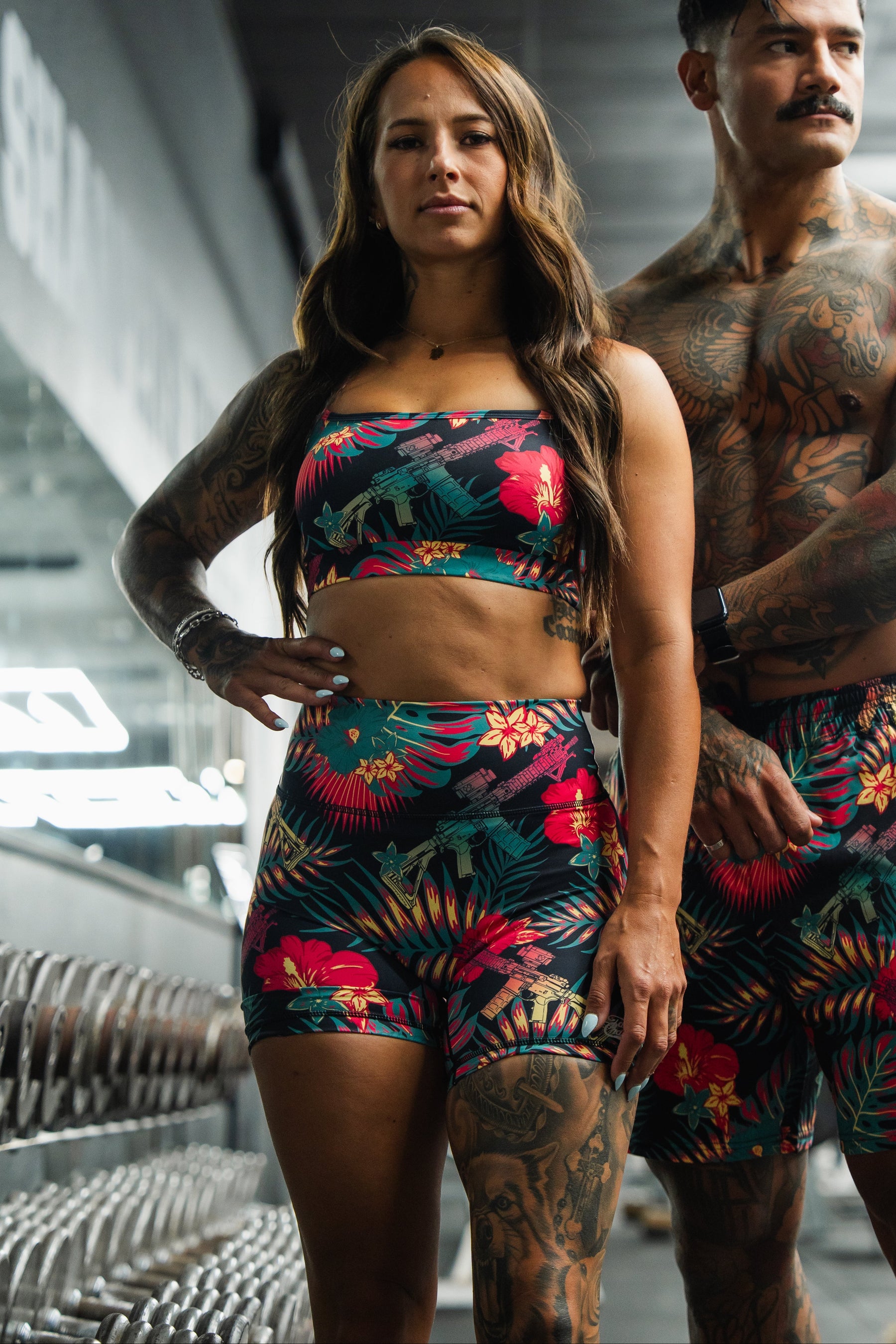 A woman stands confidently in a gym wearing the Tango Charlie Apparel Cross Back Sports Bra in the Tropical Assassin print, while a man stands behind her, both with tattoos and serious expressions, dressed in matching tropical-inspired athletic outfits.