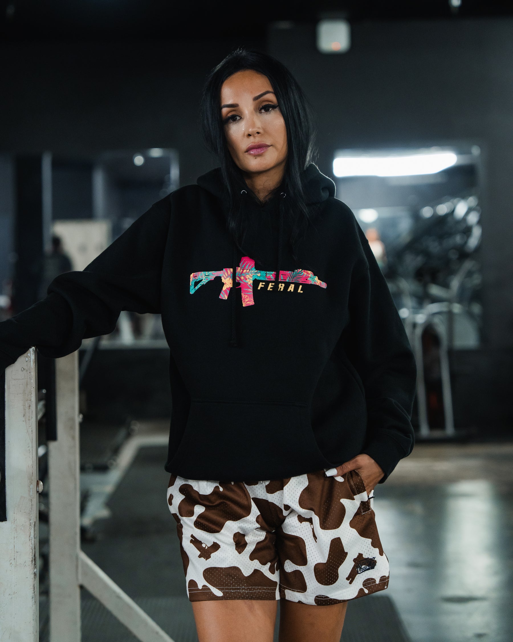 A person wearing a Tango Charlie Apparel Feral - Premium Hoodie with a floral rifle graphic and FERAL text paired with cow-print shorts, embodying radical authenticity.