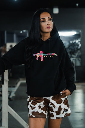 A person wearing a Tango Charlie Apparel Feral - Premium Hoodie with a tropical-print rifle graphic and cow-pattern athletic shorts, captured with feral, unfiltered observation.