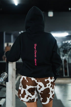 A back view of a person wearing a black Feral - Premium Hoodie with pink vertical text and cow-print athletic shorts at the gym, captured with feral, unfiltered observation.