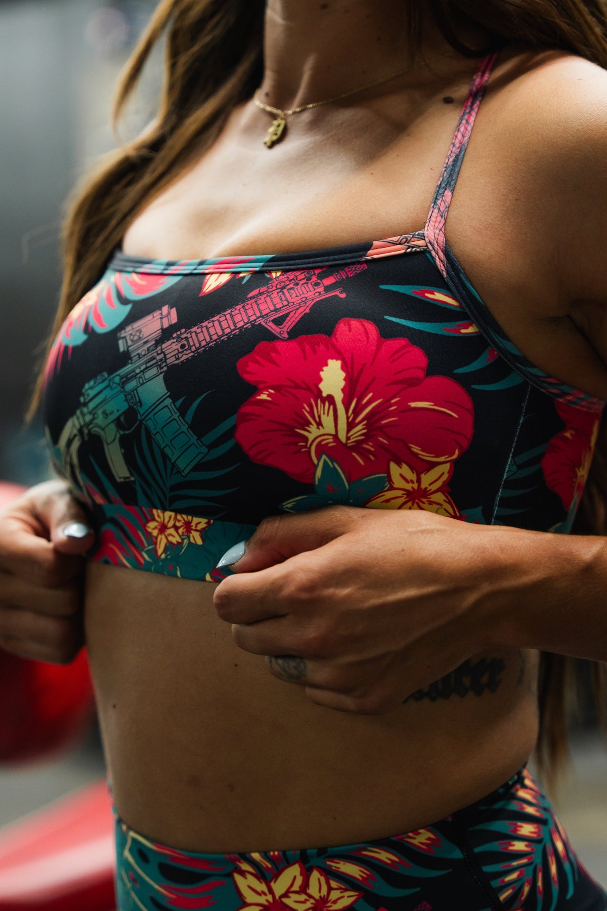 A woman adjusts the Tango Charlie Apparel Cross Back Sports Bra | Tropical Assassin, which features vibrant pink and yellow hibiscus flowers with green leaves. Only her torso and hands are visible.