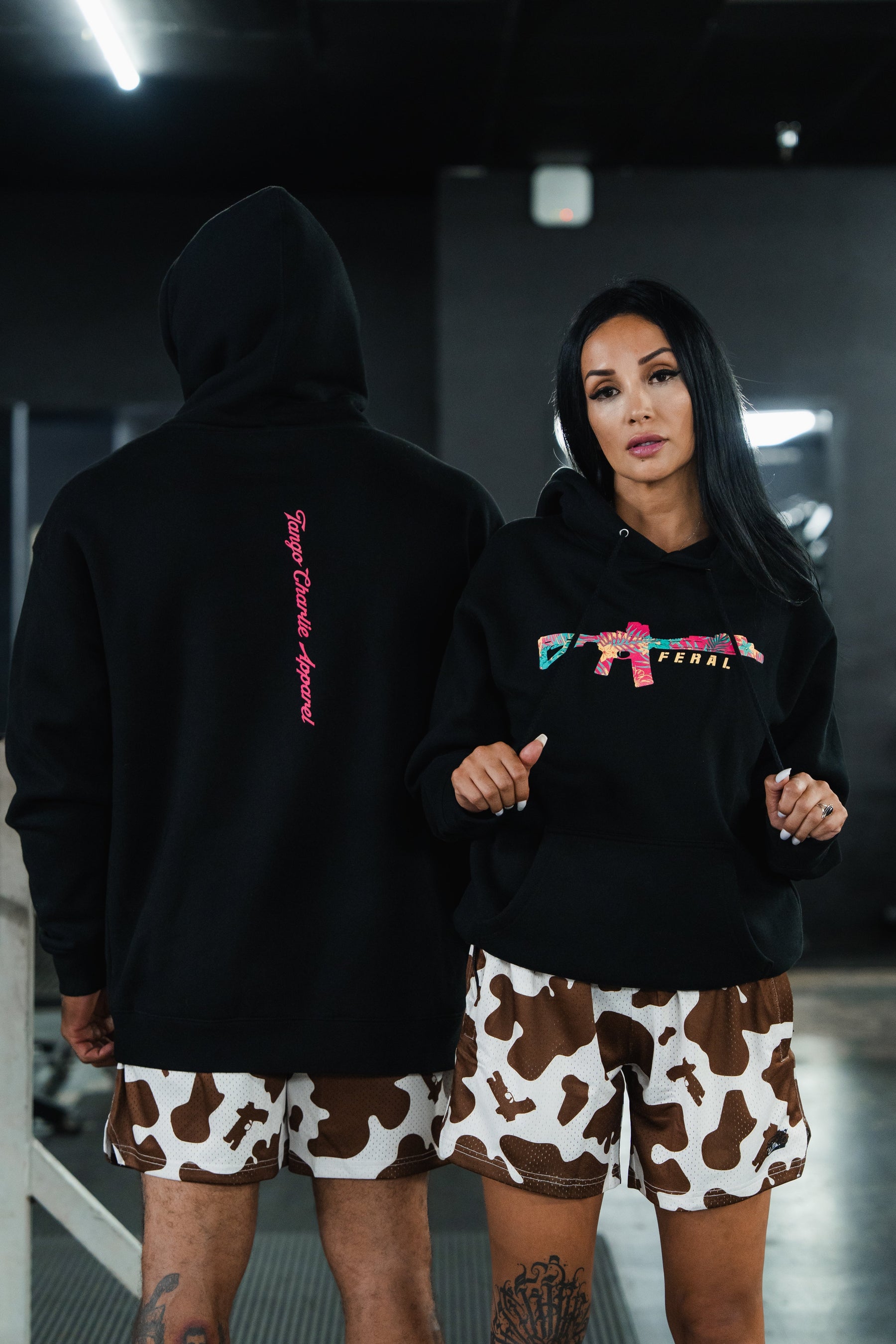 A man and a woman wearing the Feral - Premium Hoodie, featuring black fabric with pink text and a colorful rifle graphic, paired with cow-print shorts in a display of feral, unfiltered observation.