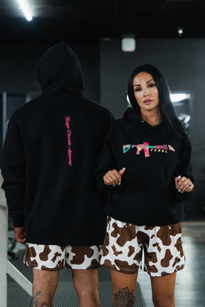 A man and a woman wearing the Feral - Premium Hoodie, featuring black fabric with pink text and a colorful rifle graphic, paired with cow-print shorts in a display of feral, unfiltered observation.