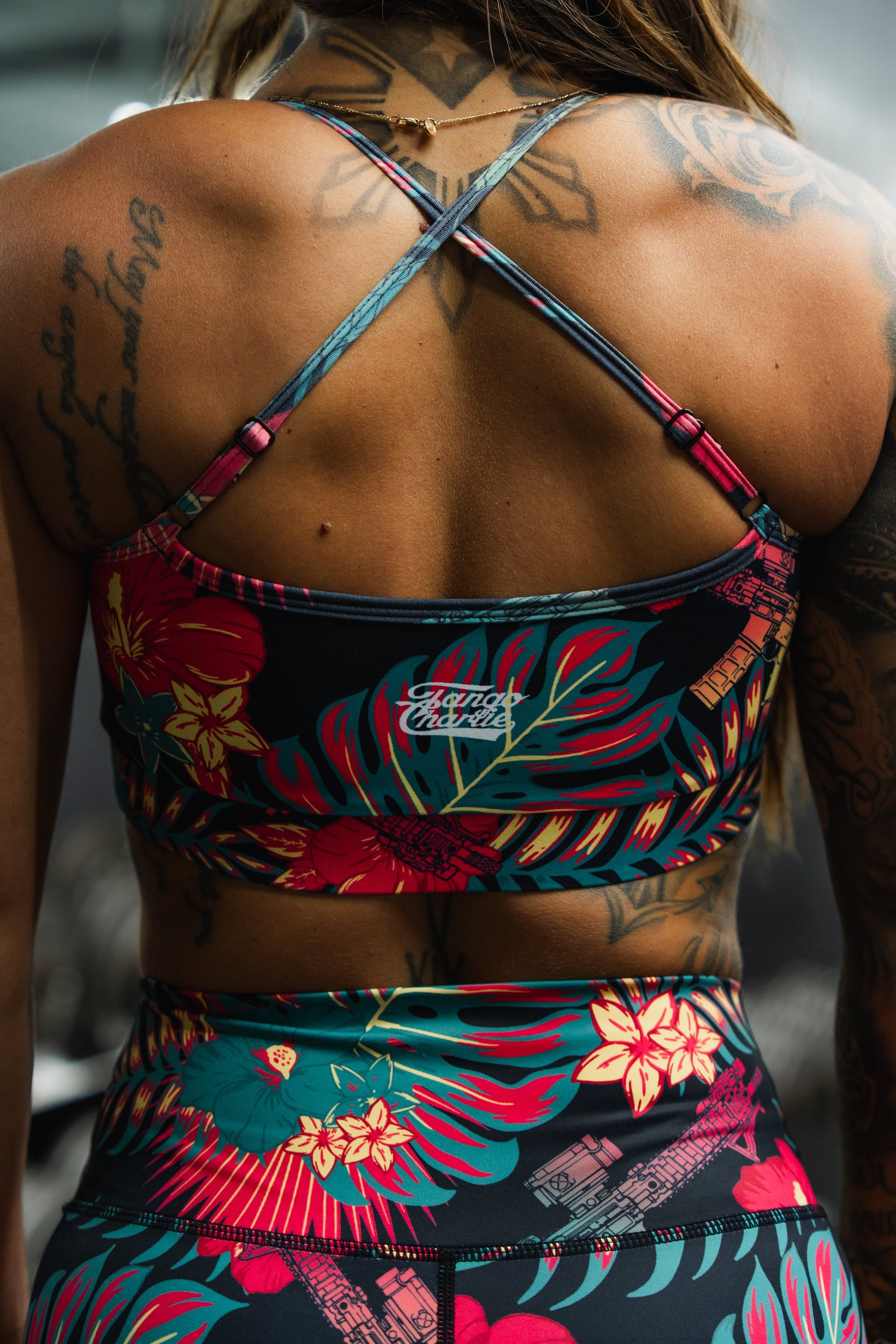 A tattooed woman wears the Tango Charlie Apparel Cross Back Sports Bra | Tropical Assassin and matching leggings. The crisscross straps and bold tropical print accentuate her upper back while providing stylish, medium support.
