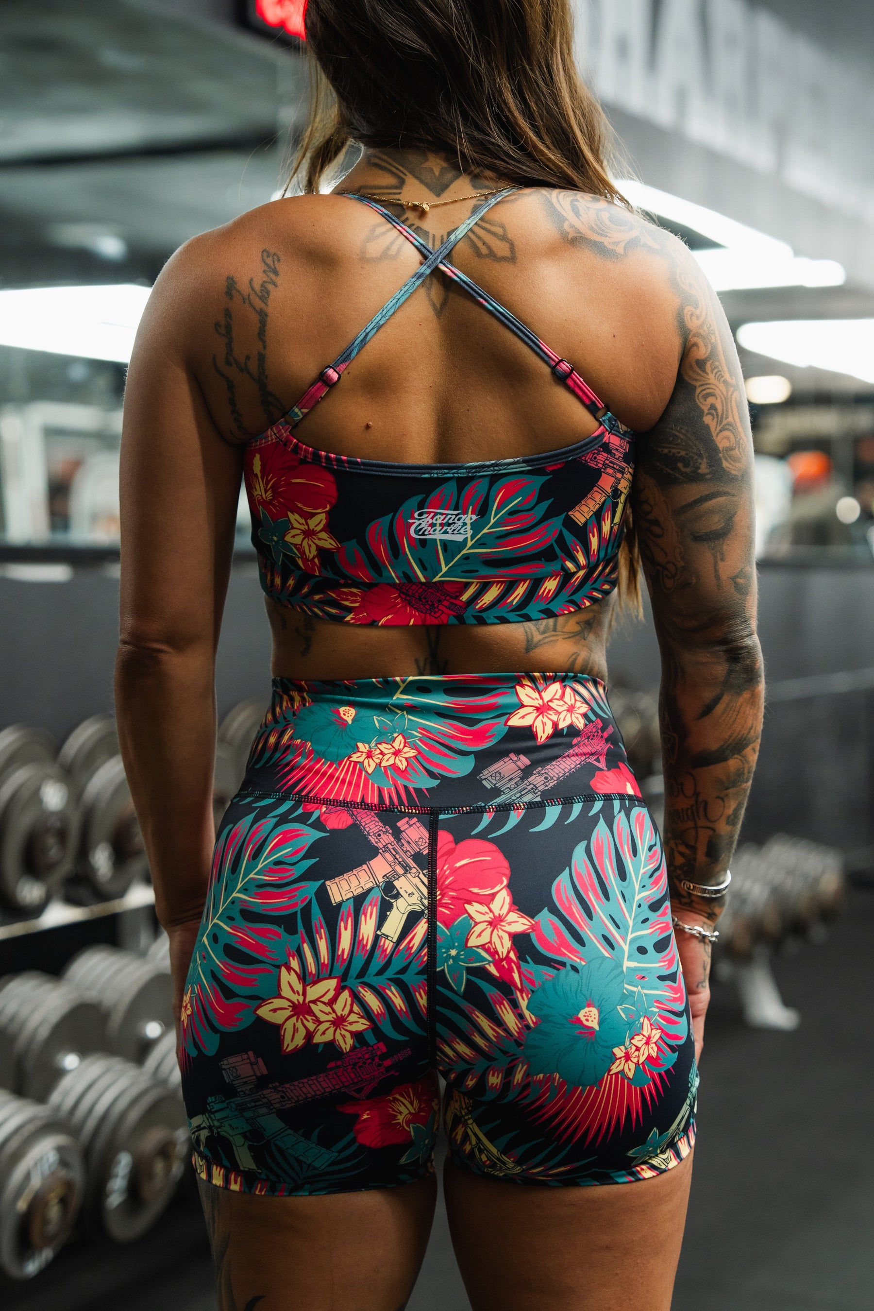 A tattooed woman wearing Tango Charlie Apparel's Women's High-Waisted Seamless Front Compression Training Shorts | Tropical Assassin stands in a gym, facing away from the camera, with dumbbells and equipment visible in the background.