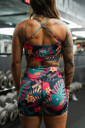 A tattooed woman wearing Tango Charlie Apparel's Women's High-Waisted Seamless Front Compression Training Shorts | Tropical Assassin stands in a gym, facing away from the camera, with dumbbells and equipment visible in the background.