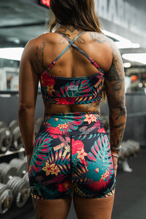 A tattooed woman stands in a gym wearing the Tango Charlie Apparel Cross Back Sports Bra | Tropical Assassin and shorts, facing away from the camera with gym weights and mirrors in the background.