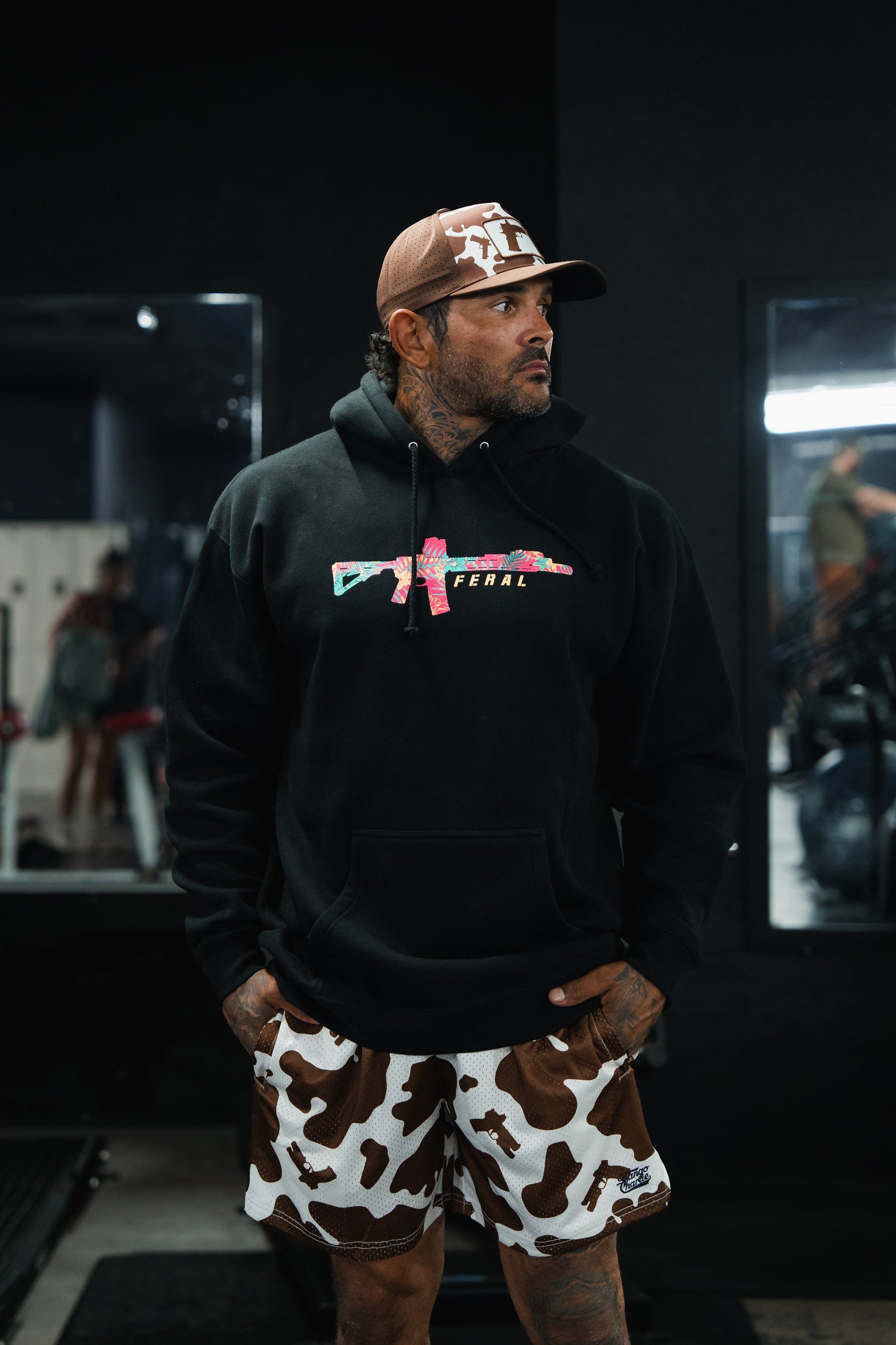 A man wears a black Feral - Premium Hoodie with a floral-patterned rifle graphic, paired with cow-print shorts and a cap.