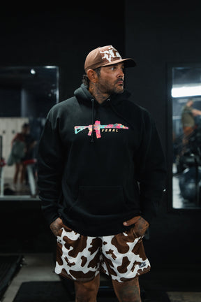 A man wears a black Feral - Premium Hoodie with a floral-patterned rifle graphic, paired with cow-print shorts and a cap.