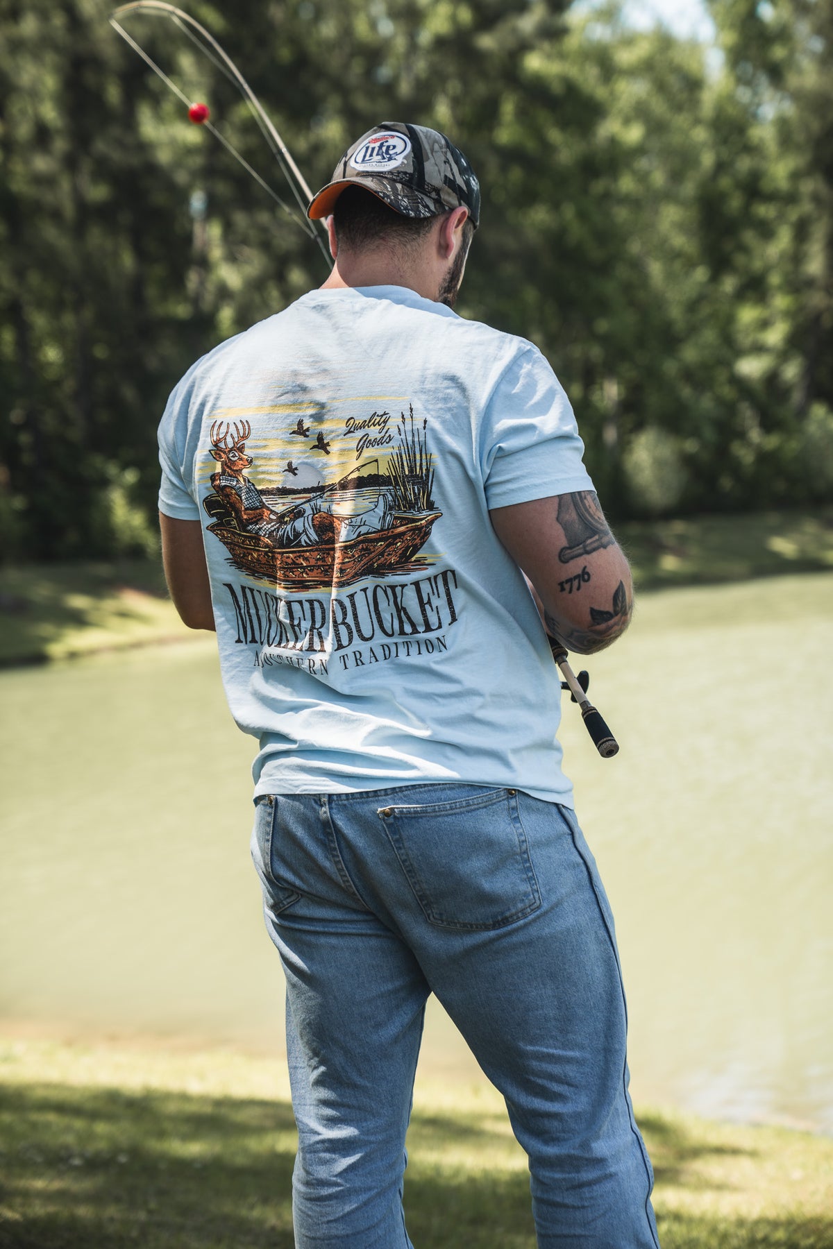 Wearing his Mucker Bucket Buck Dreamin’ Premium Tee, a man stands by the pond casting his line, surrounded by lush greenery as he enjoys a peaceful day outdoors.