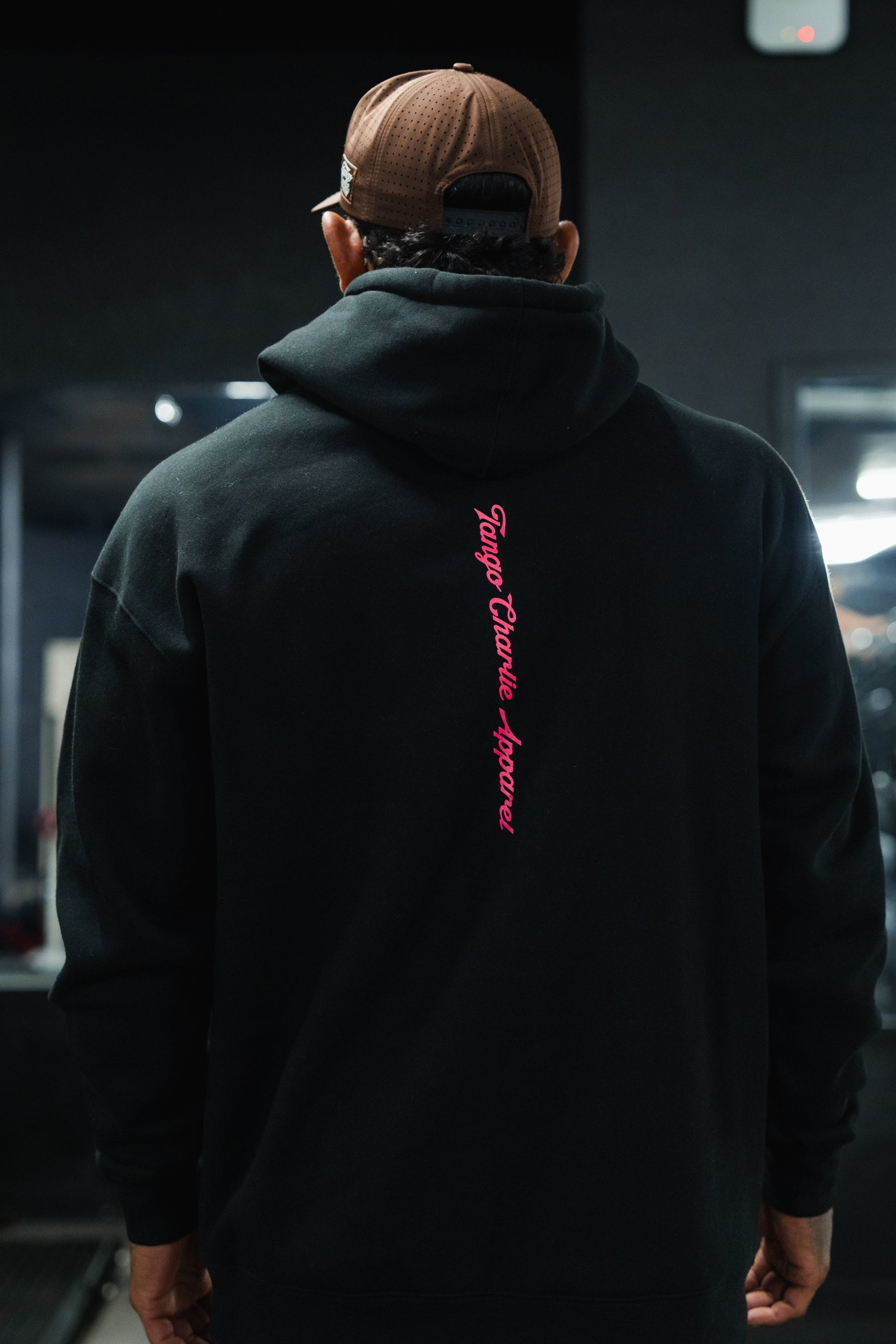 Back view of a person wearing a brown baseball cap and a black Feral - Premium Hoodie by Tango Charlie Apparel with pink vertical text, capturing a sense of radical authenticity in an unfiltered observation.