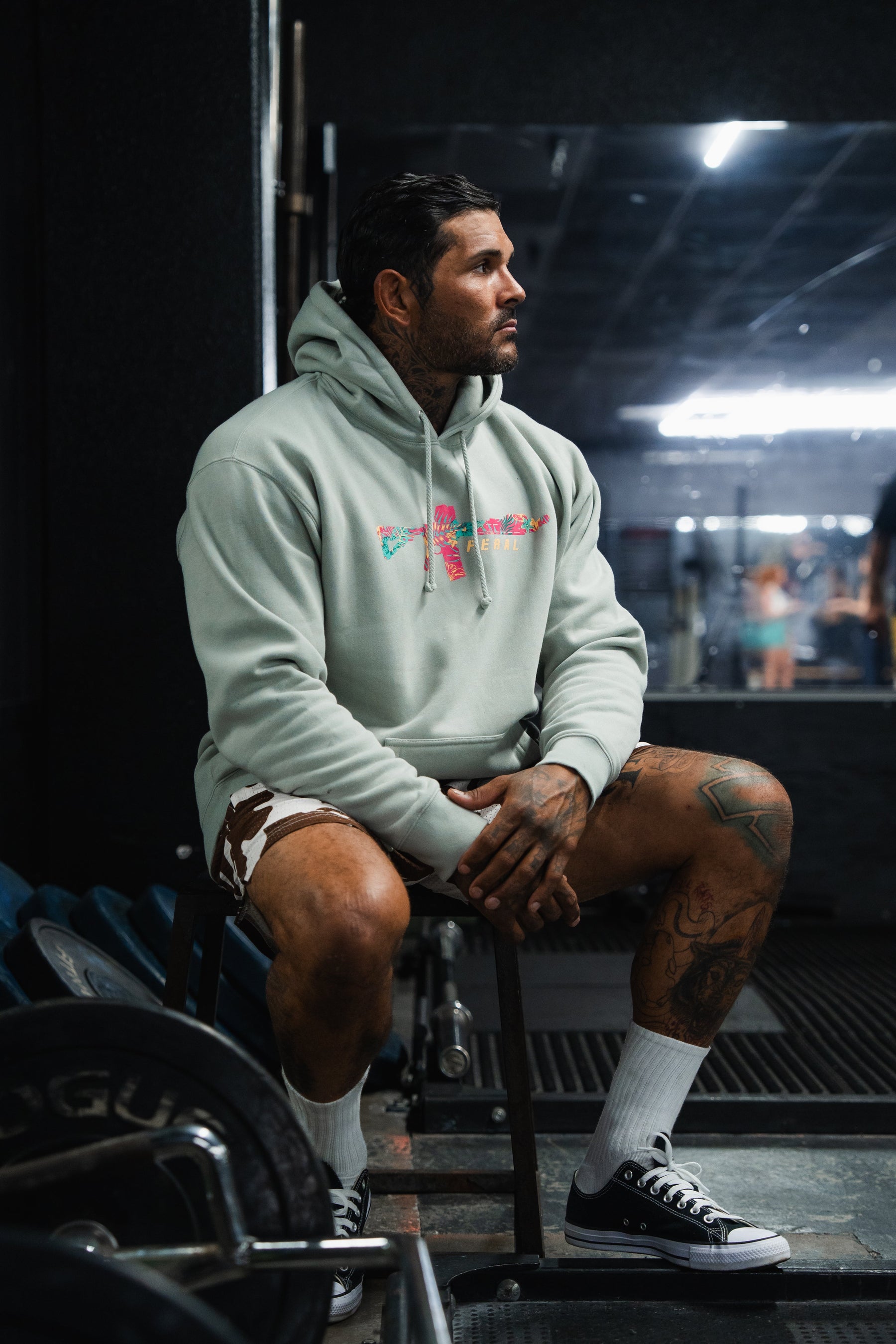 A tattooed man sits in a gym wearing a Feral - Premium Dusty Sage Hoodie with a floral rifle graphic, radiating feral, unfiltered observation.