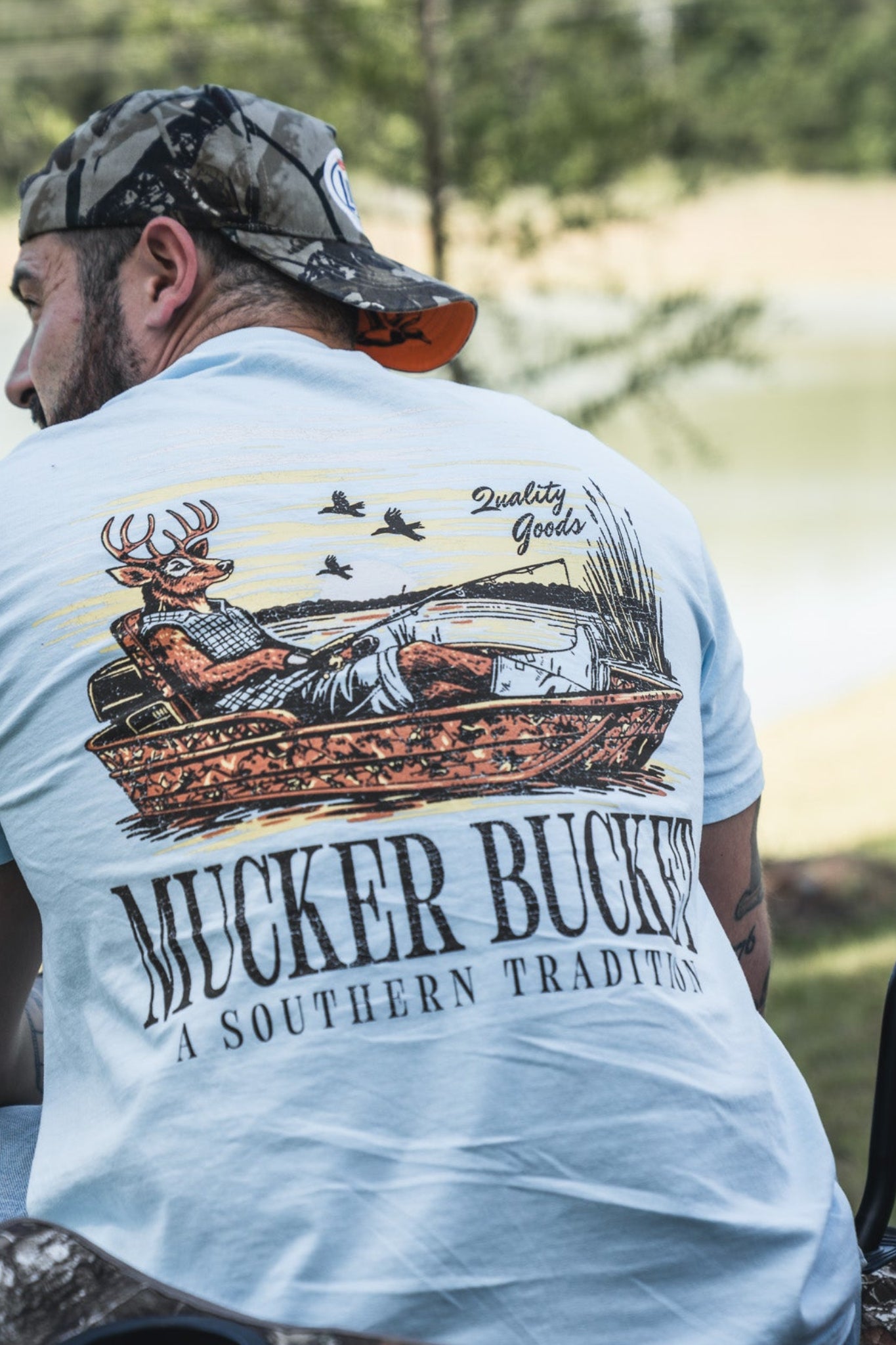 A man relaxes outdoors near a pond and trees in the Buck Dreamin' Premium Tee by Mucker Bucket, enjoying the comfort of this premium cotton fishing t-shirt featuring a deer graphic and “A Southern Tradition” slogan.