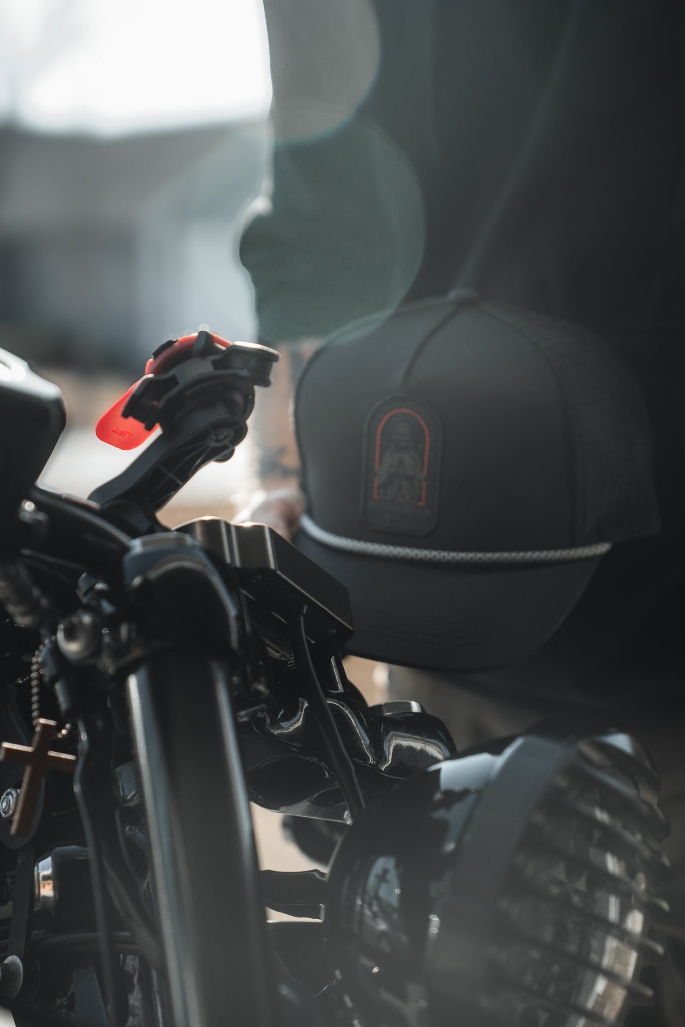 A close-up shows a motorcycle handlebar and headlight, with a person wearing the Born United Private Label Saints and Sinners Hat. The subtle saintly design on the hat catches the sunlight, creating an ethereal lens flare effect in the background.