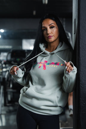 Woman wearing a Feral - Premium Dusty Sage Hoodie with a floral-print rifle graphic and the text FERAL on the chest.