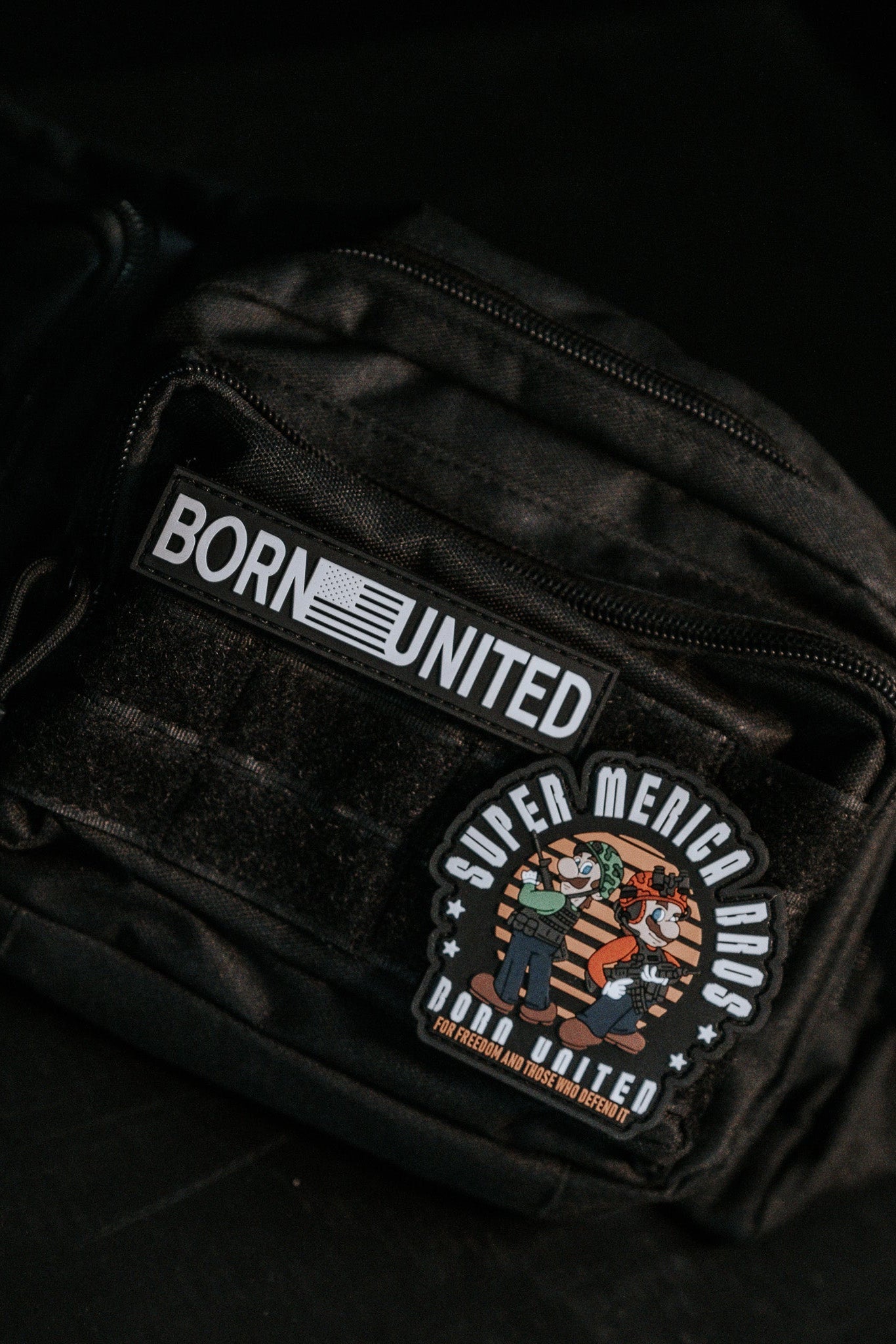 The Midnight Black Tac-Fanny-Pac by Born United Private Label features a BORN UNITED patch and a SUPER MERICA BROS. round patch with playful hero characters, adding a whimsical touch to its sleek design.
