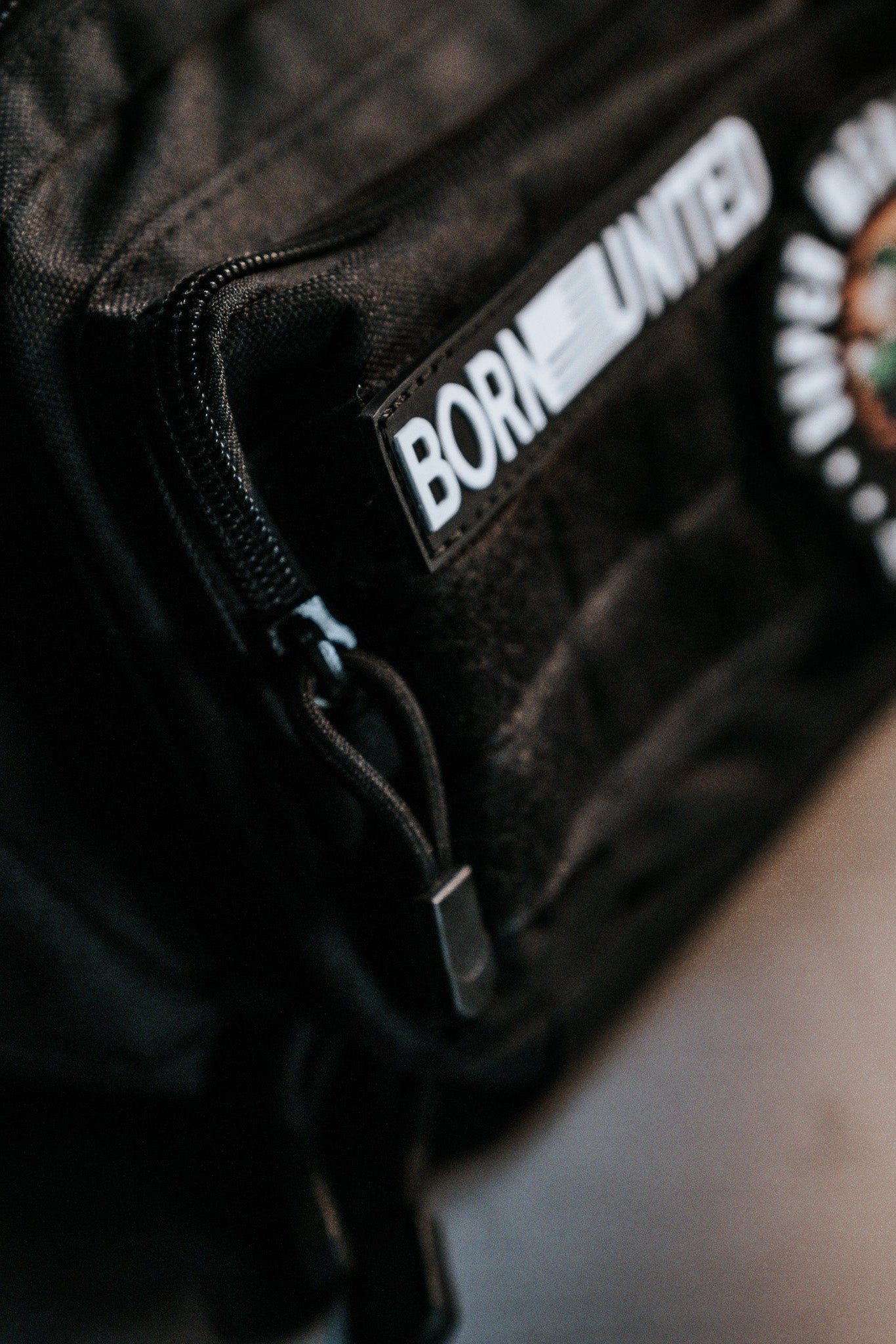 Close-up of a black Midnight Black Tac-Fanny-Pac by Born United Private Label. A patch with BORN UNITED in white letters is seen on a zippered pocket, revealing the fabric texture, while another colorful patch is partially visible.