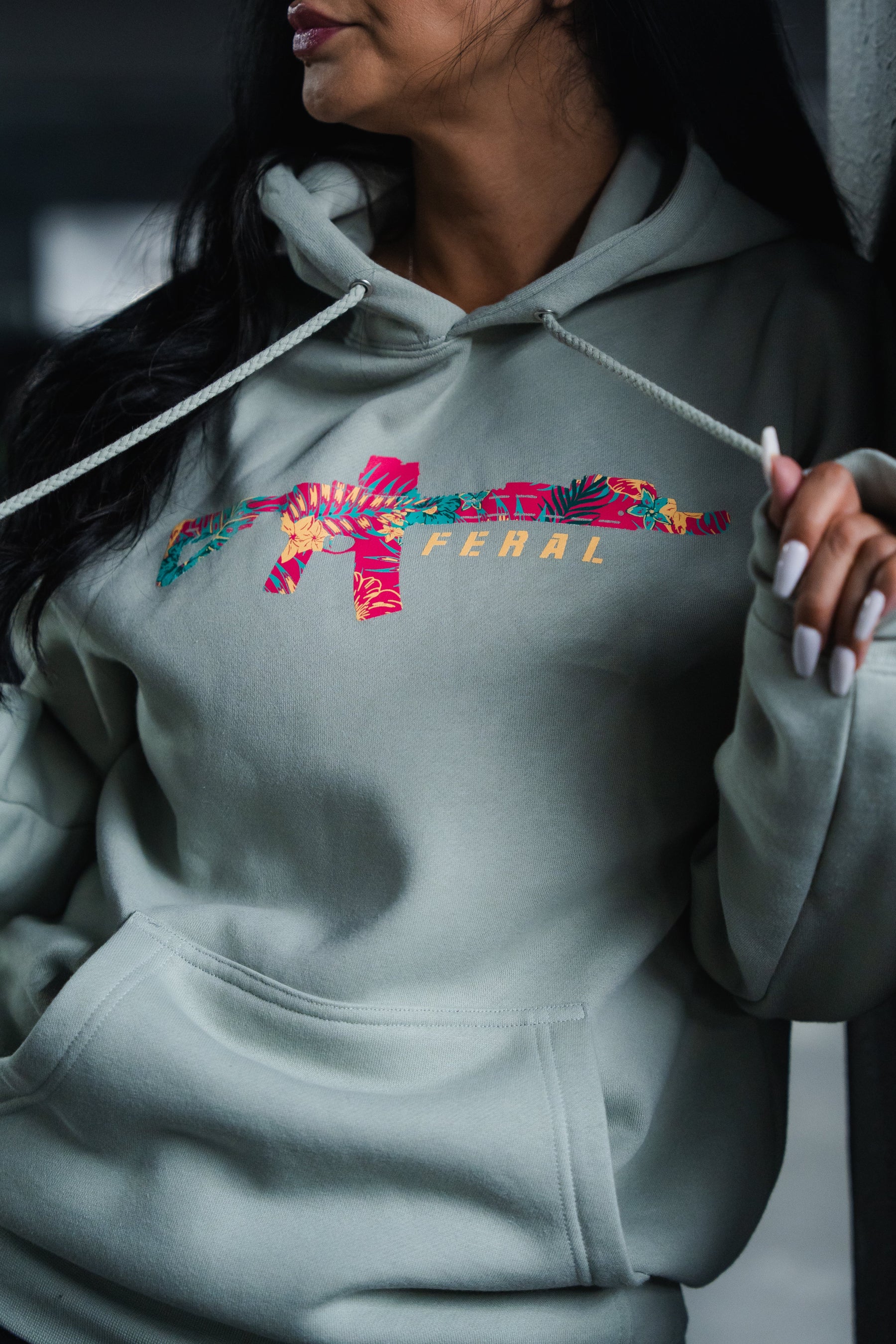 A person wears a Feral - Premium Dusty Sage Hoodie featuring a graphic of a rifle in a tropical floral print with the word FERAL printed beneath it.