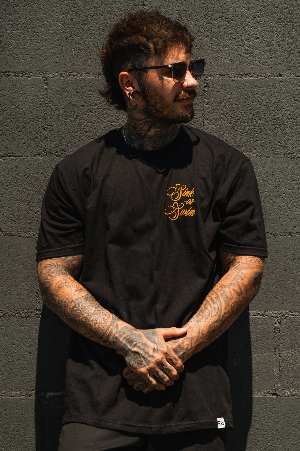 A tattooed person in sunglasses and the Relentless Betrayal Sink Or Swim Kraken T-Shirt, featuring gold lettering on fine jersey cotton, stands against a gray brick wall looking sideways with hands clasped in front.