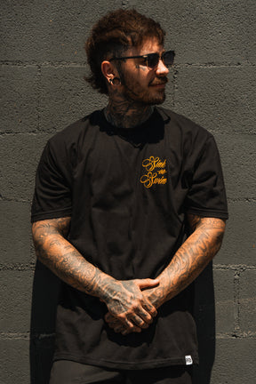 A tattooed person in sunglasses and the Relentless Betrayal Sink Or Swim Kraken T-Shirt, featuring gold lettering on fine jersey cotton, stands against a gray brick wall looking sideways with hands clasped in front.