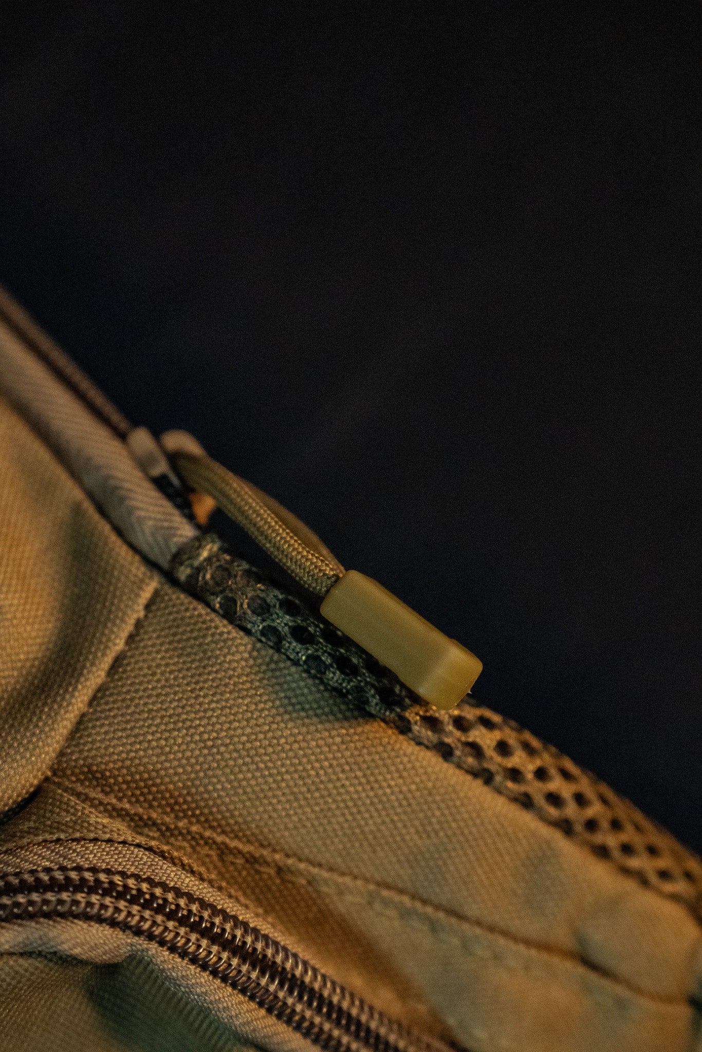 Close-up of a tan fabric zipper on the Coyote Tac-Fanny-Pac by Born United Private Label. The zipper pull is connected to a green cord, with textured fabric. A dark background highlights the zipper focus.