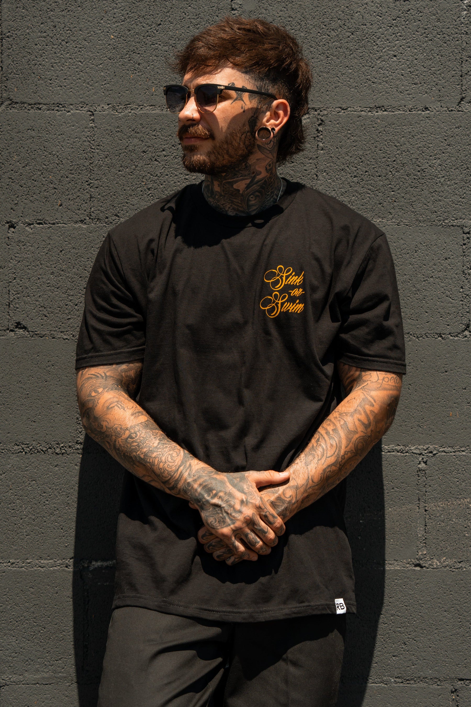 A tattooed man with dark hair and sunglasses stands against a gray brick wall, wearing the Relentless Betrayal Sink Or Swim Kraken T-Shirt with yellow script on the chest, looking to the side with hands clasped in front.