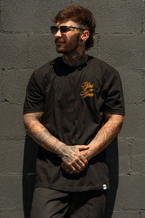 A tattooed man with dark hair and sunglasses stands against a gray brick wall, wearing the Relentless Betrayal Sink Or Swim Kraken T-Shirt with yellow script on the chest, looking to the side with hands clasped in front.