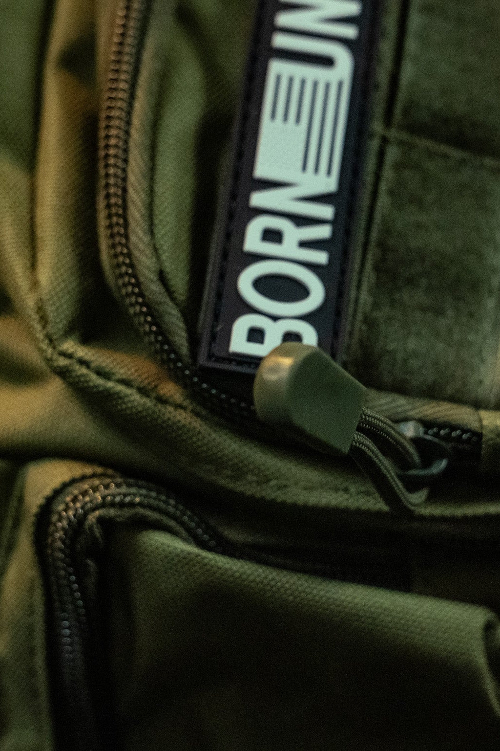 A close-up of the OD Green Tac-Fanny-Pac by Born United Private Label shows a visible zipper and patch with BORN, followed by an unreadable section. Dim lighting highlights the fabric texture and zipper pull, enhancing its rugged style.