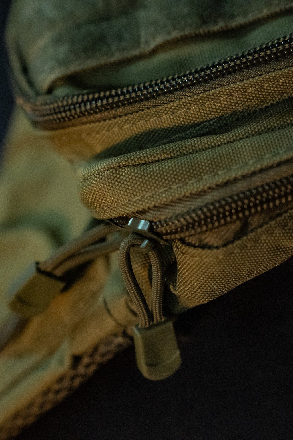 The OD Green Tac-Fanny-Pac by Born United Private Label is shown in a close-up, highlighting its tactical canvas texture, stitching, and durable zipper pulls. The low-light setting emphasizes its rugged design, resembling an oversized fanny pack.