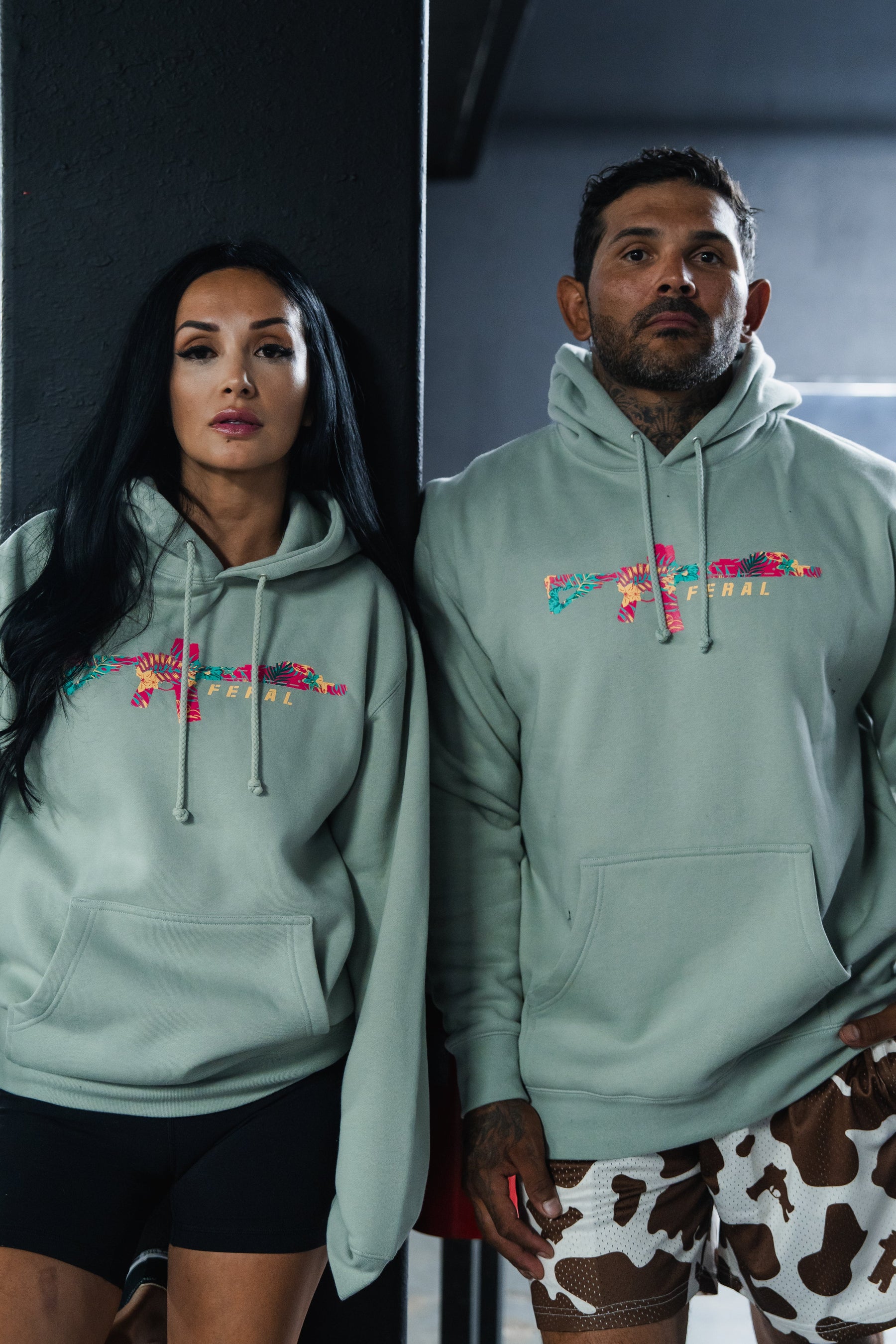 A man and woman wearing matching Feral - Premium Dusty Sage Hoodies with a floral rifle graphic, embodying radical authenticity.