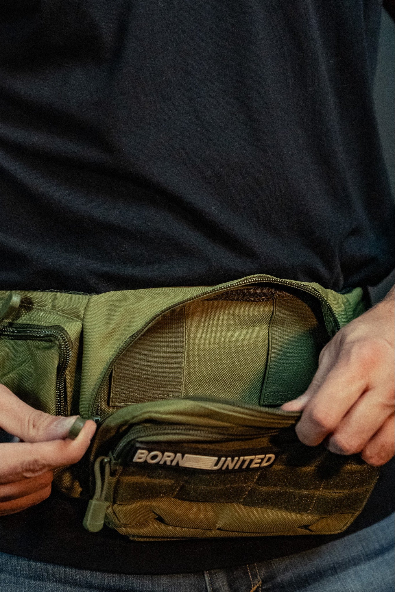 A person in a black shirt adjusts a green tactical pouch resembling a fanny pack. The OD Green Tac-Fanny-Pac by Born United Private Label features multiple compartments, with BORN UNITED partially visible on the front pocket.