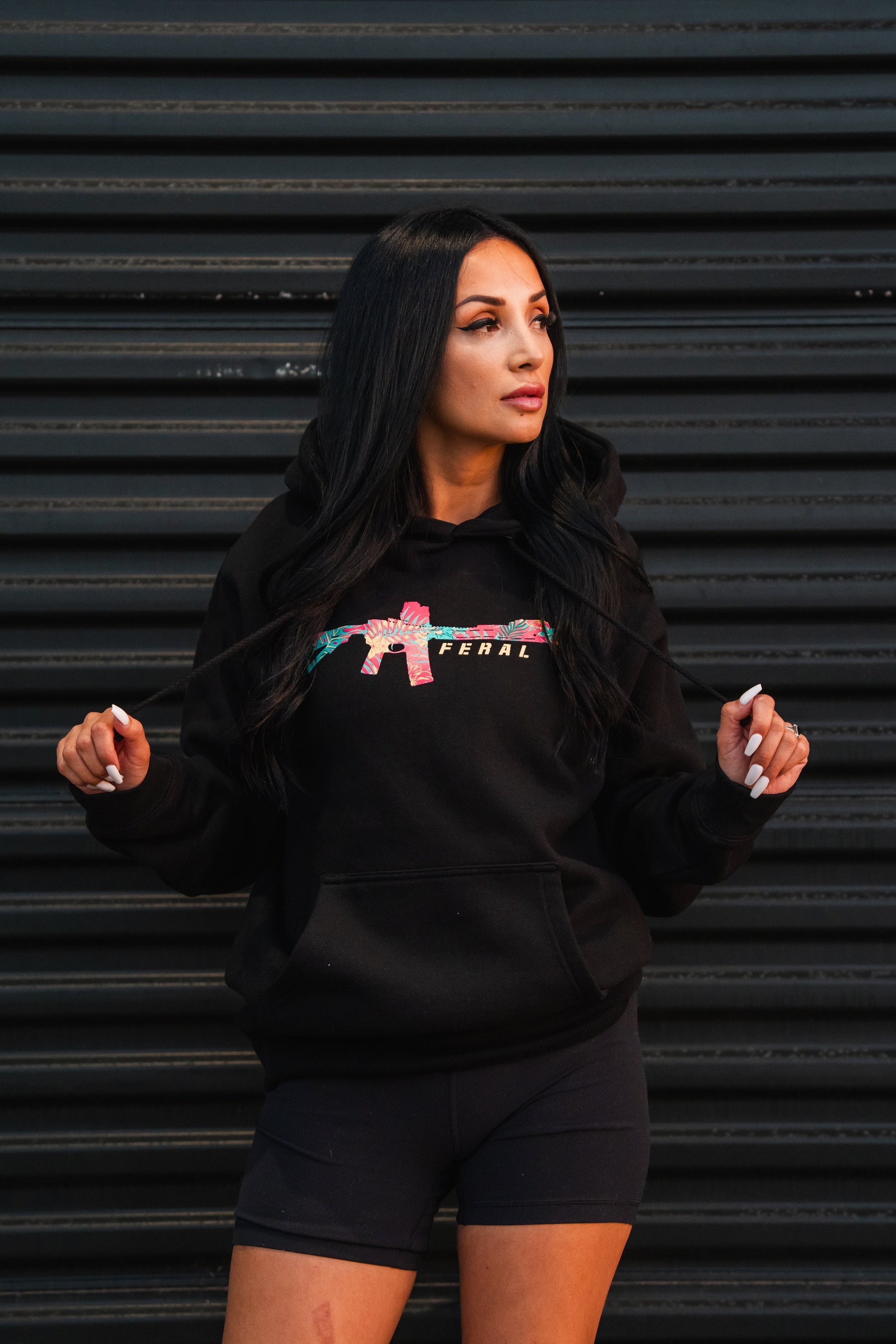 A woman wearing a black Feral - Premium Hoodie featuring a tropical-print rifle graphic and the word FERAL on the chest.