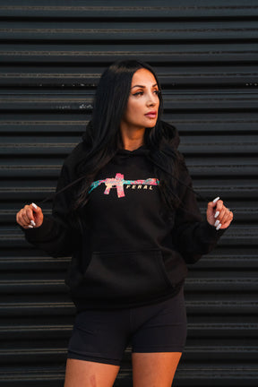 A woman wearing a black Feral - Premium Hoodie featuring a tropical-print rifle graphic and the word FERAL on the chest.