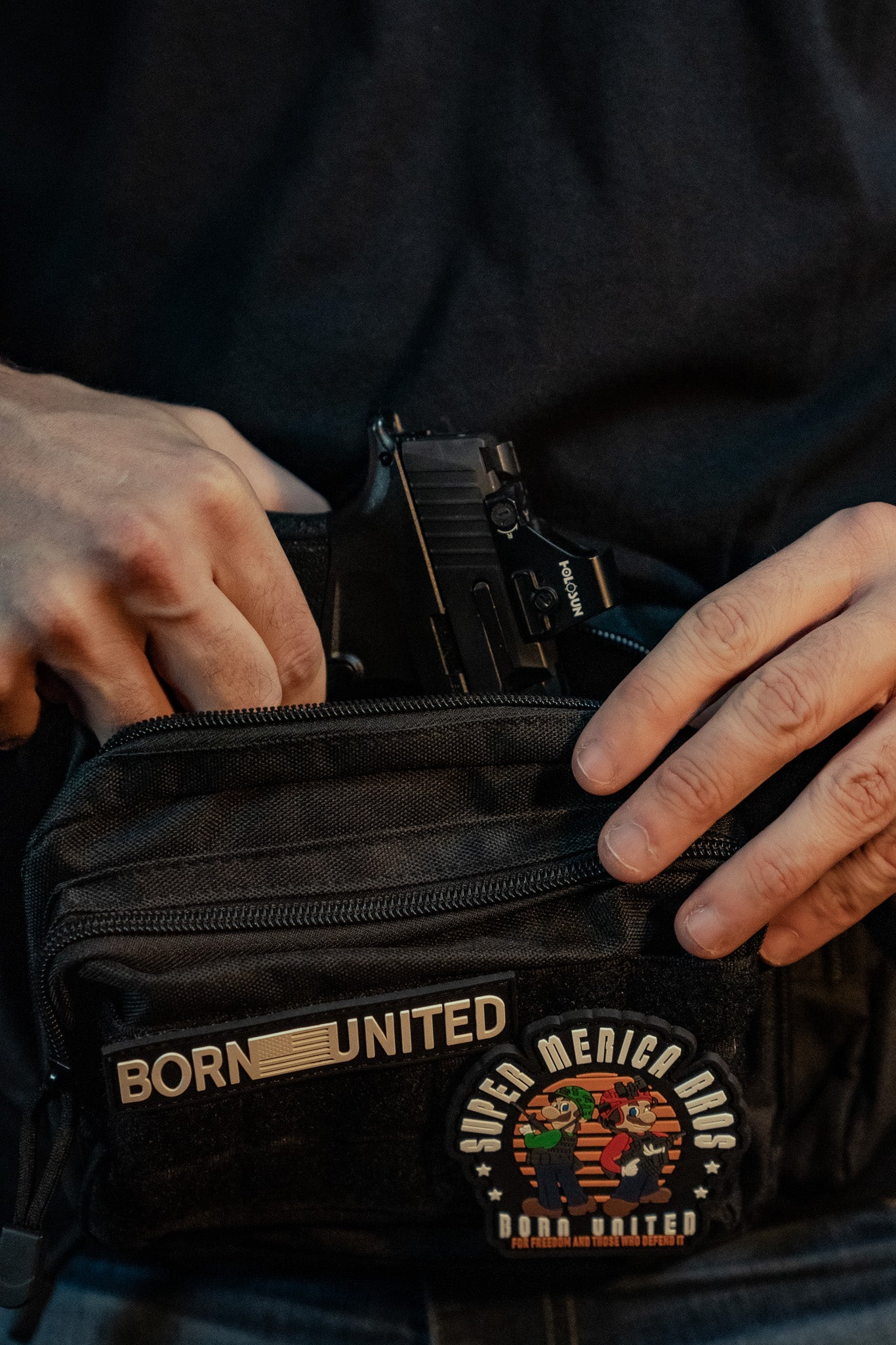 A person in a black shirt discreetly tucks a handgun into the Midnight Black Tac-Fanny-Pac by Born United Private Label, featuring two patches: one with BORN UNITED and another showcasing a colorful SUPER MERICA RAC design.