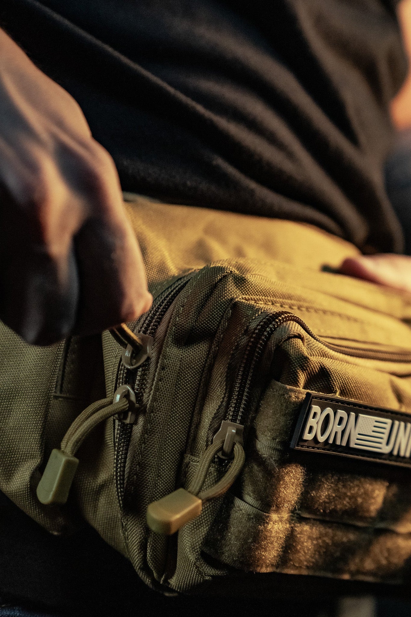 A person wearing a dark shirt adjusts their Coyote Tac-Fanny-Pac from Born United Private Label, which is designed in tactical beige with yellow zipper pulls and showcases a BORN UNITED patch.