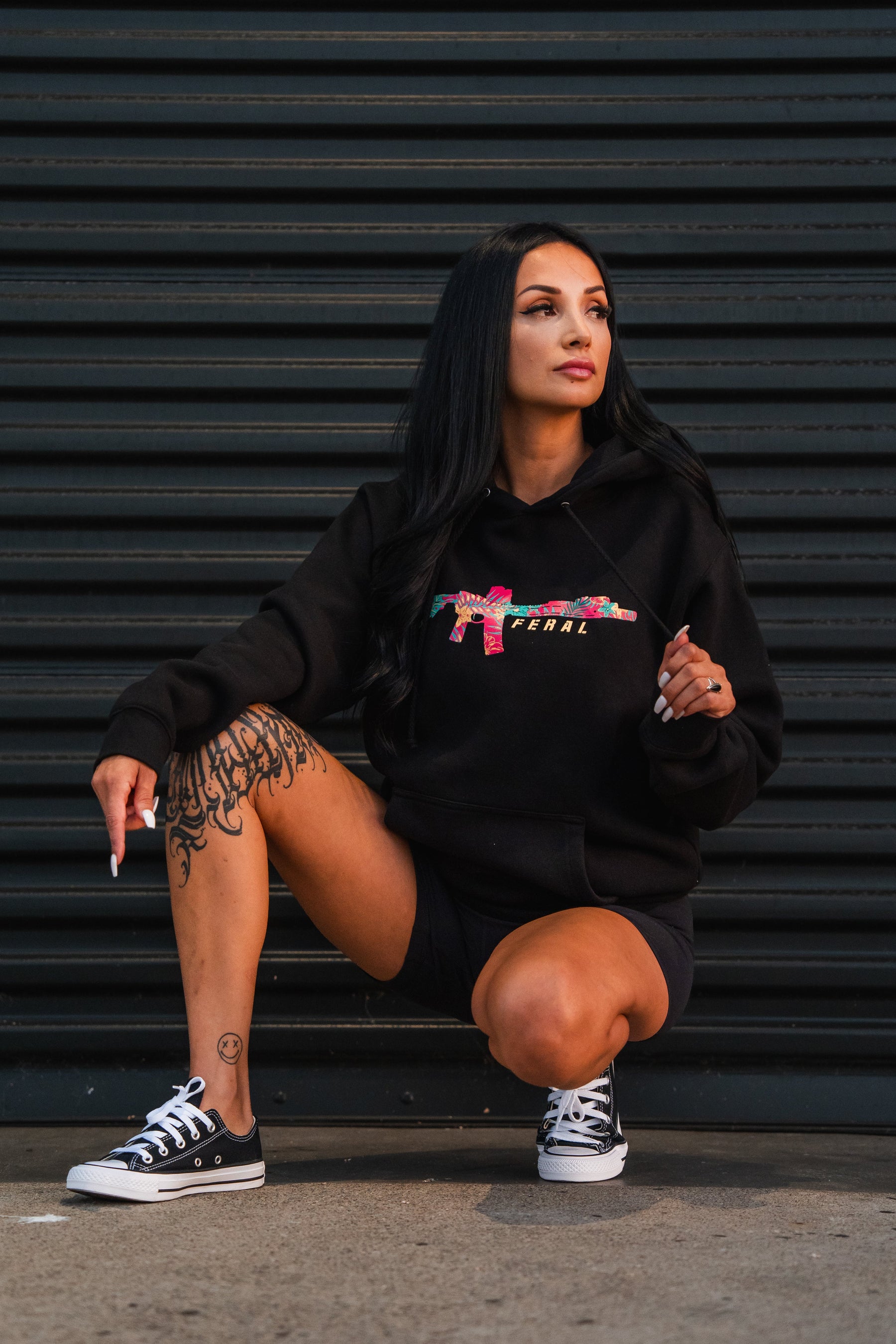 A woman crouches against a black corrugated metal wall wearing a Tango Charlie Apparel Feral - Premium Hoodie, which features a floral rifle graphic and FERAL printed on the chest.