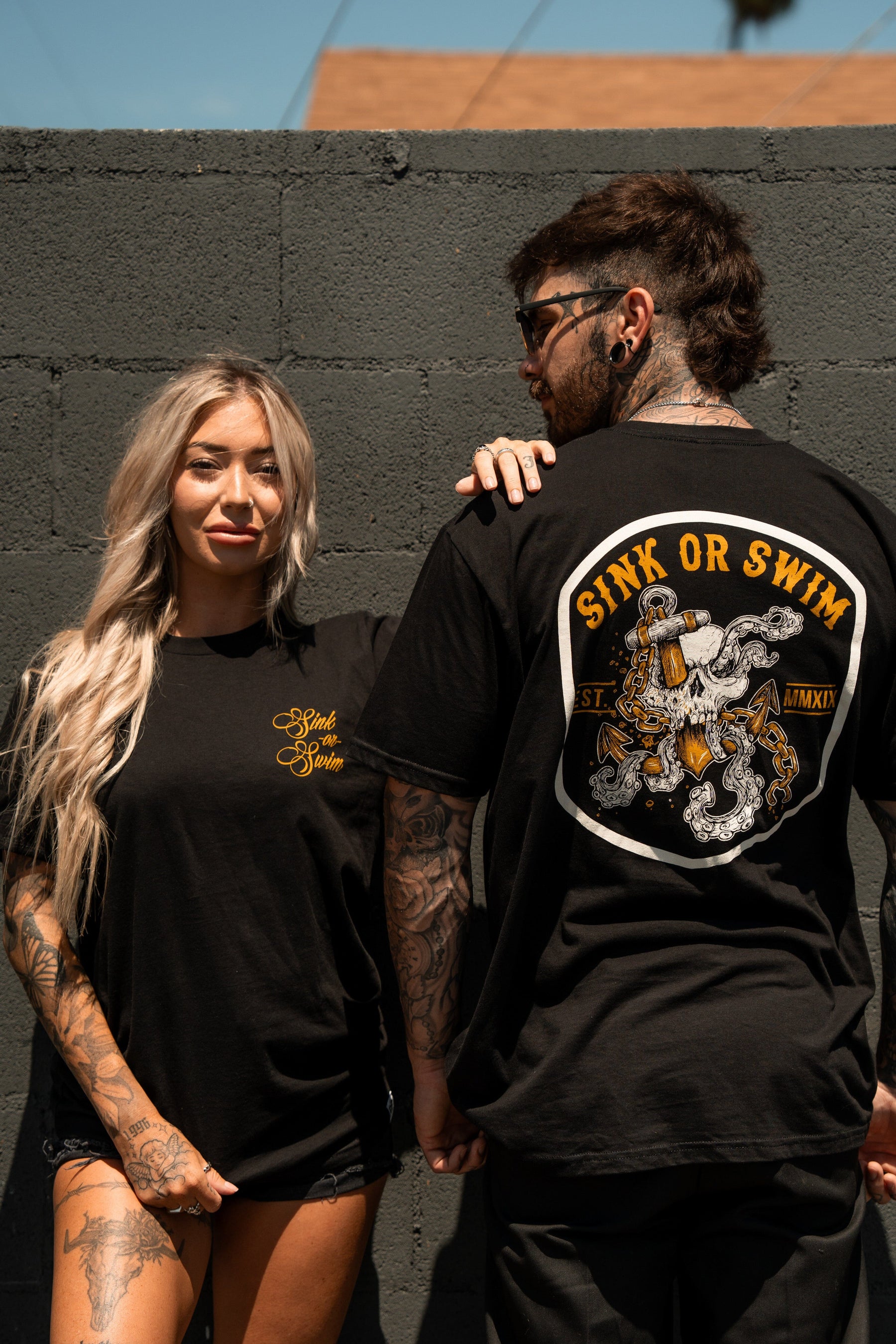 A tattooed woman faces forward in a black Relentless Betrayal Sink Or Swim Kraken T-Shirt with yellow script, while a man stands beside her, showing the motivational back graphic of his fine jersey cotton shirt.