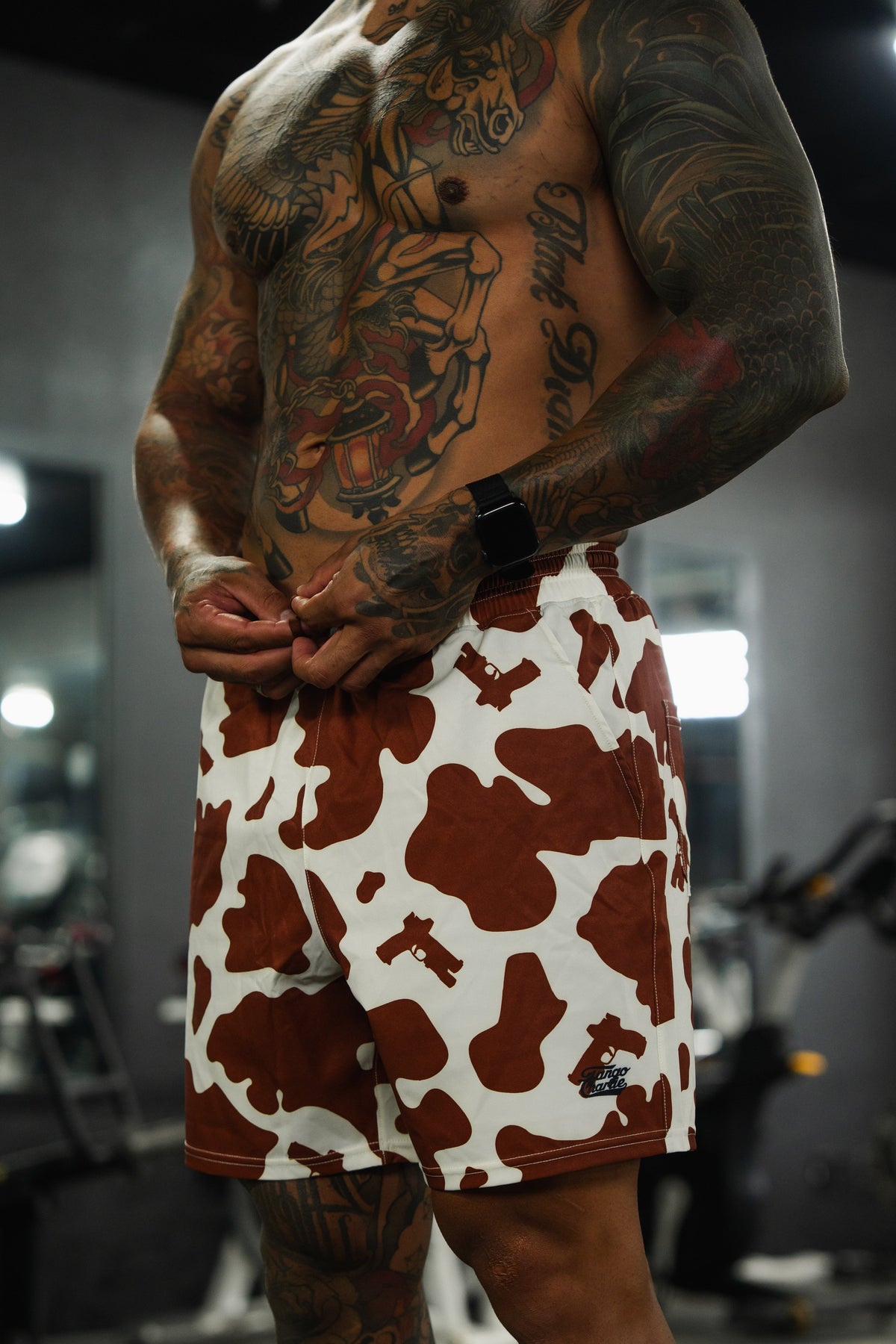A muscular, tattooed person stands in a gym wearing Tango Charlie Apparel's Men's Performance Training Shorts | 5.5"- Moo Moo Pew, holding the waistband; blurred gym equipment and mirrors are visible in the background.