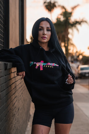 Woman wearing a black Feral - Premium Hoodie with a colorful, patterned rifle graphic and the word FERAL on the chest, capturing a sense of radical authenticity.