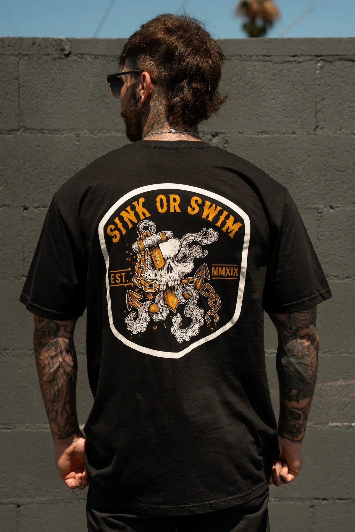 A tattooed individual faces a gray brick wall wearing the Relentless Betrayal Sink Or Swim Kraken T-Shirt, which showcases a graphic octopus with chains and anchor plus "SINK OR SWIM EST. MMXIX" text on the back.