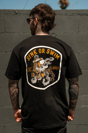 A tattooed individual faces a gray brick wall wearing the Relentless Betrayal Sink Or Swim Kraken T-Shirt, which showcases a graphic octopus with chains and anchor plus "SINK OR SWIM EST. MMXIX" text on the back.
