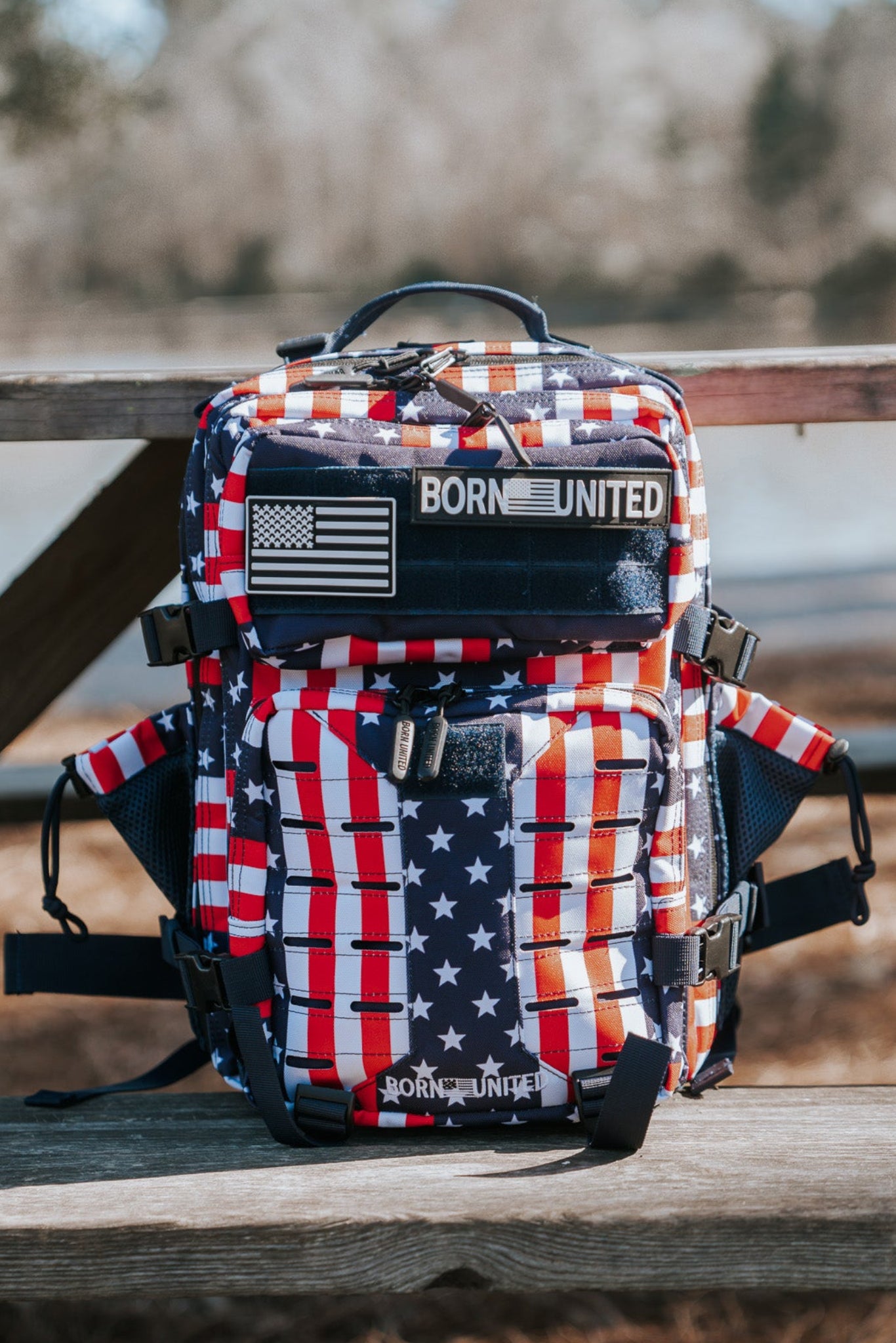 The Ultimate Patriot Special Edition Tac-Pac - 25L by Born United Private Label, with stars and stripes design, sits on a wooden picnic table outdoors, flaunting a U.S. flag patch. Trees and a shimmering lake are visible in the background.