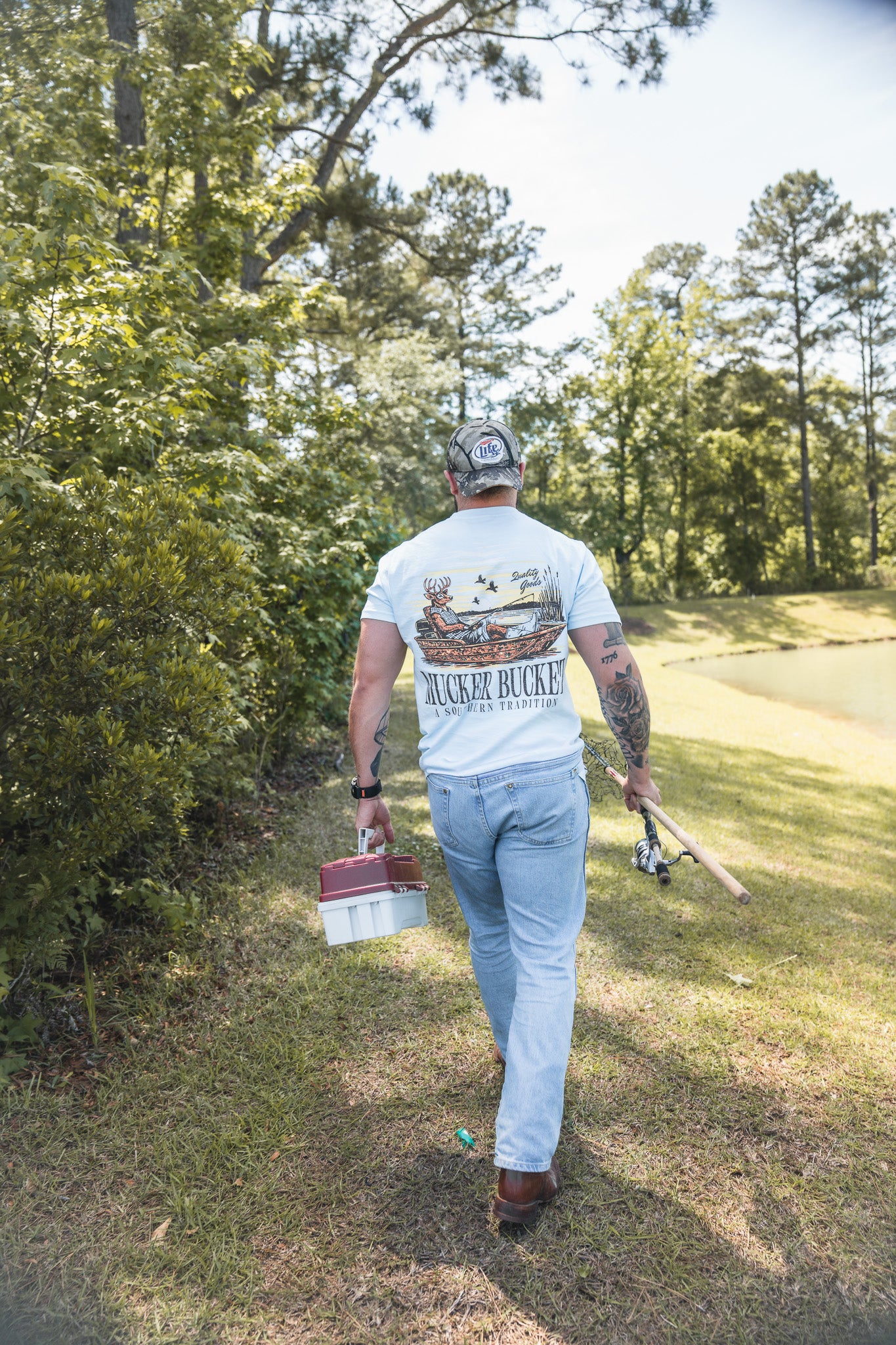 A man in a Mucker Bucket Buck Dreamin' Premium Tee, jeans, and a cap walks away on a grassy path with fishing gear, surrounded by trees under a clear sky.