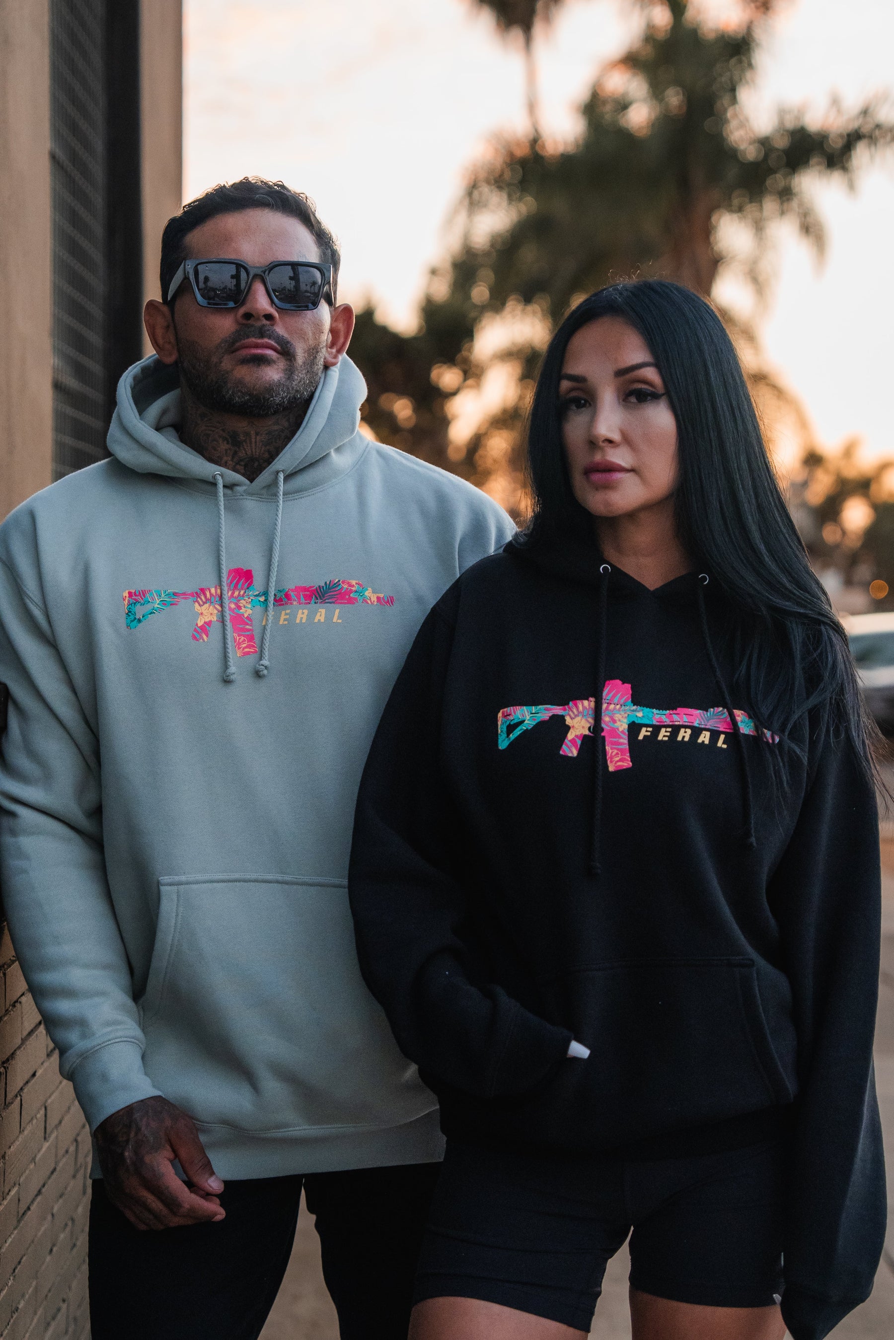 A man and woman wearing dusty sage Feral - Premium Dusty Sage Hoodies with a floral rifle graphic and the word FERAL across the chest.