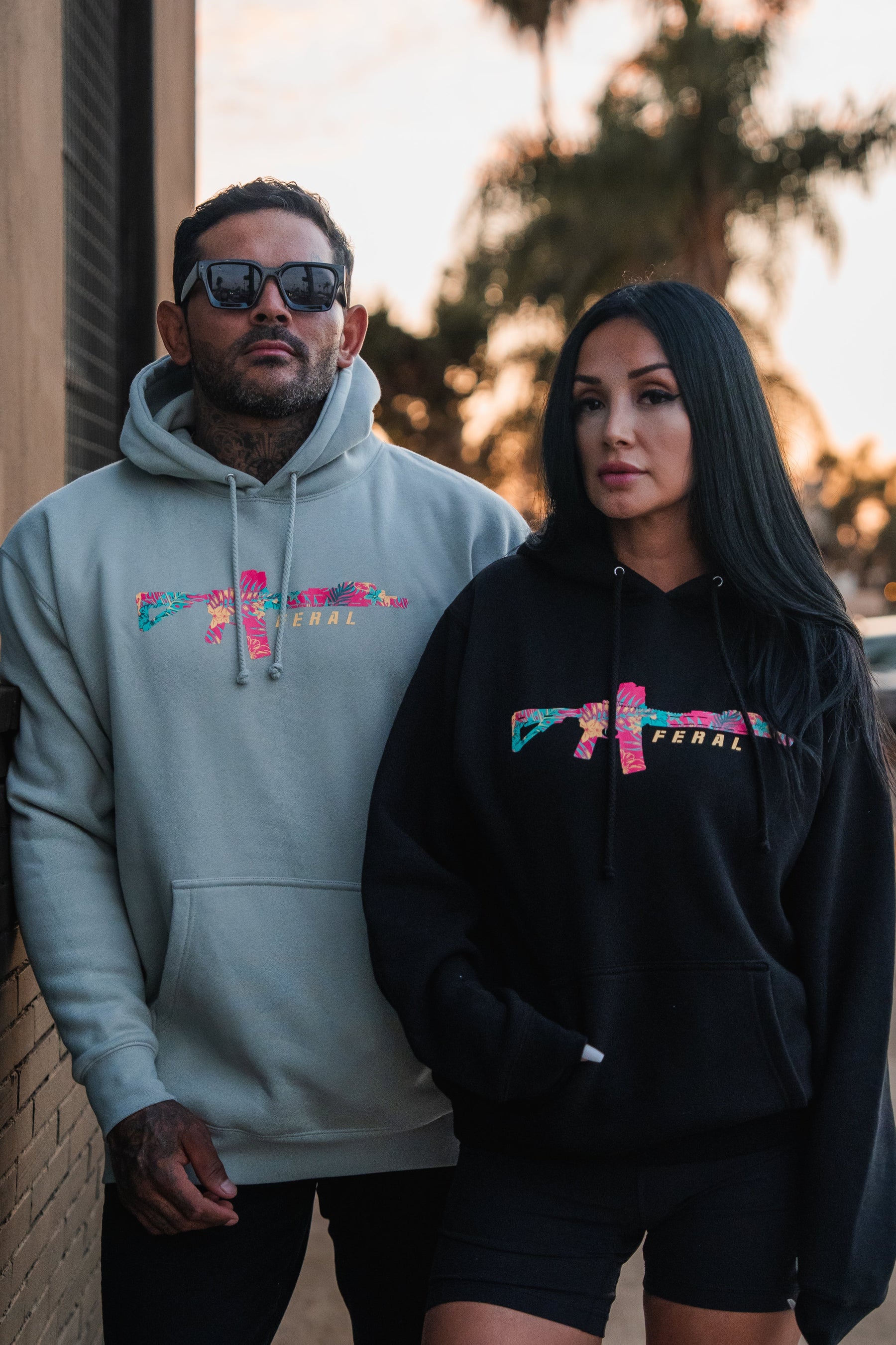 A man and woman stand side-by-side in matching Feral - Premium Hoodies, each featuring a tropical floral graphic of a rifle and the word FERAL.