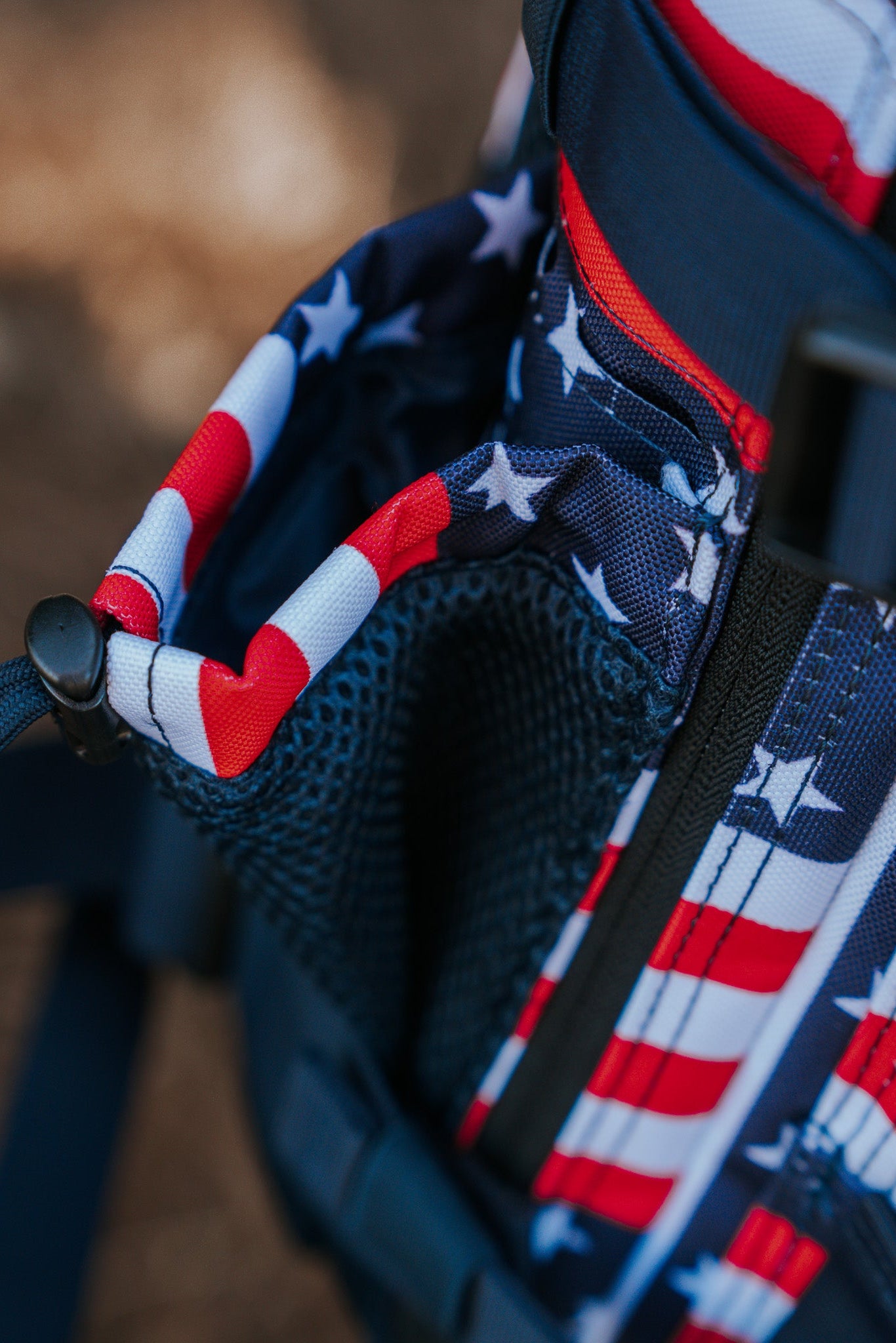 A close-up of the Born United Private Labels Ultimate Patriot Special Edition Tac-Pac - 25L, featuring an American flag design with red/white stripes and white stars on a blue background. The mesh side pocket is accented by a sleek black zipper and strap.
