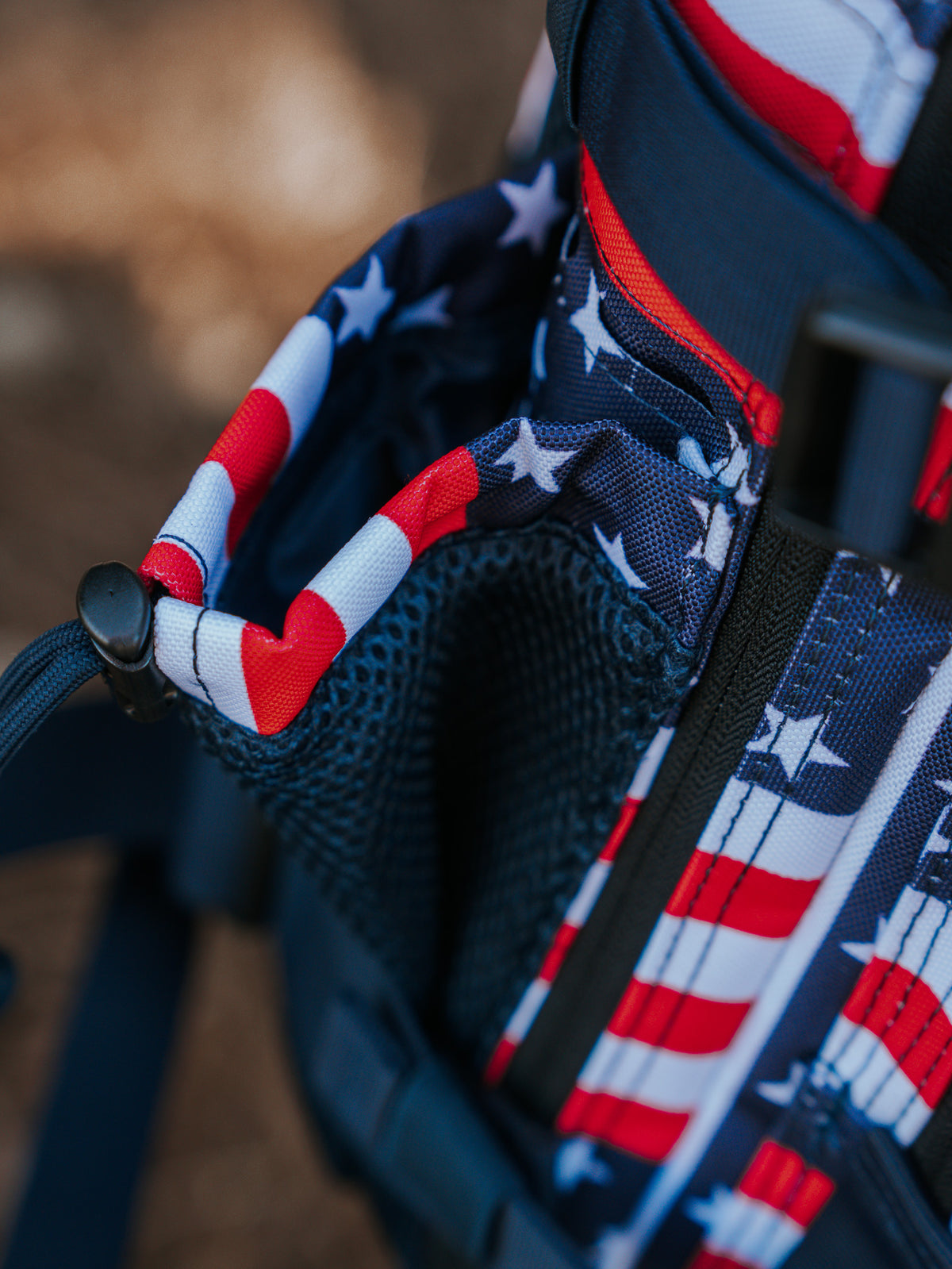 Close-up of the Born United Special Edition 25L Premium Tac-Pac Bundle backpack featuring red and white stripes with white stars on a blue background, resembling the American flag. The image shows a side pocket with black mesh, BU Patches embellishments, and a buckle strap.