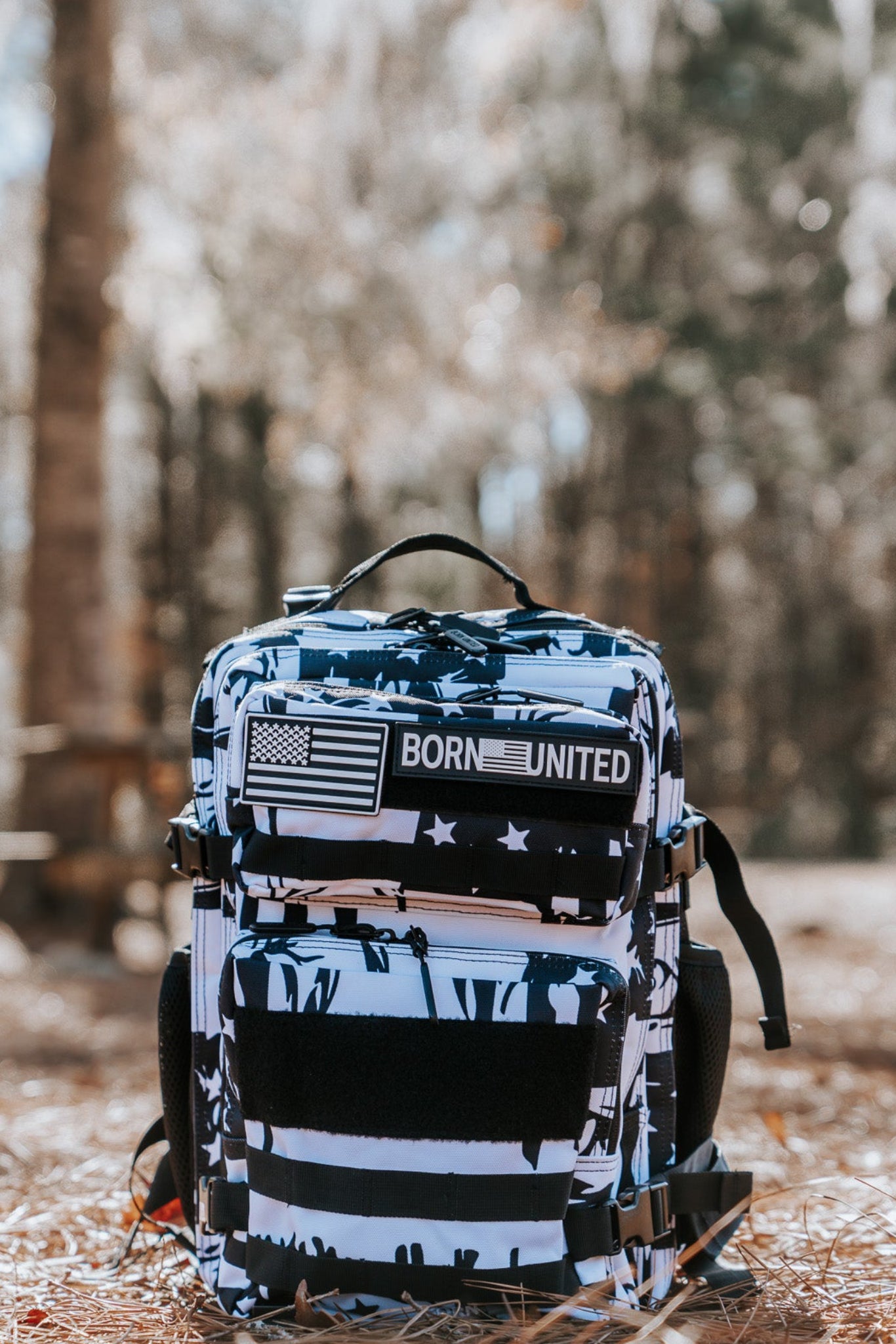 The American Stealth Special Edition Tac-Pac - 25L by Born United Private Label sits proudly on pine needles, featuring a multi-colored design with a Born United patch and American flag. A serene forest scene with blurred trees and picnic tables enriches the backdrop.